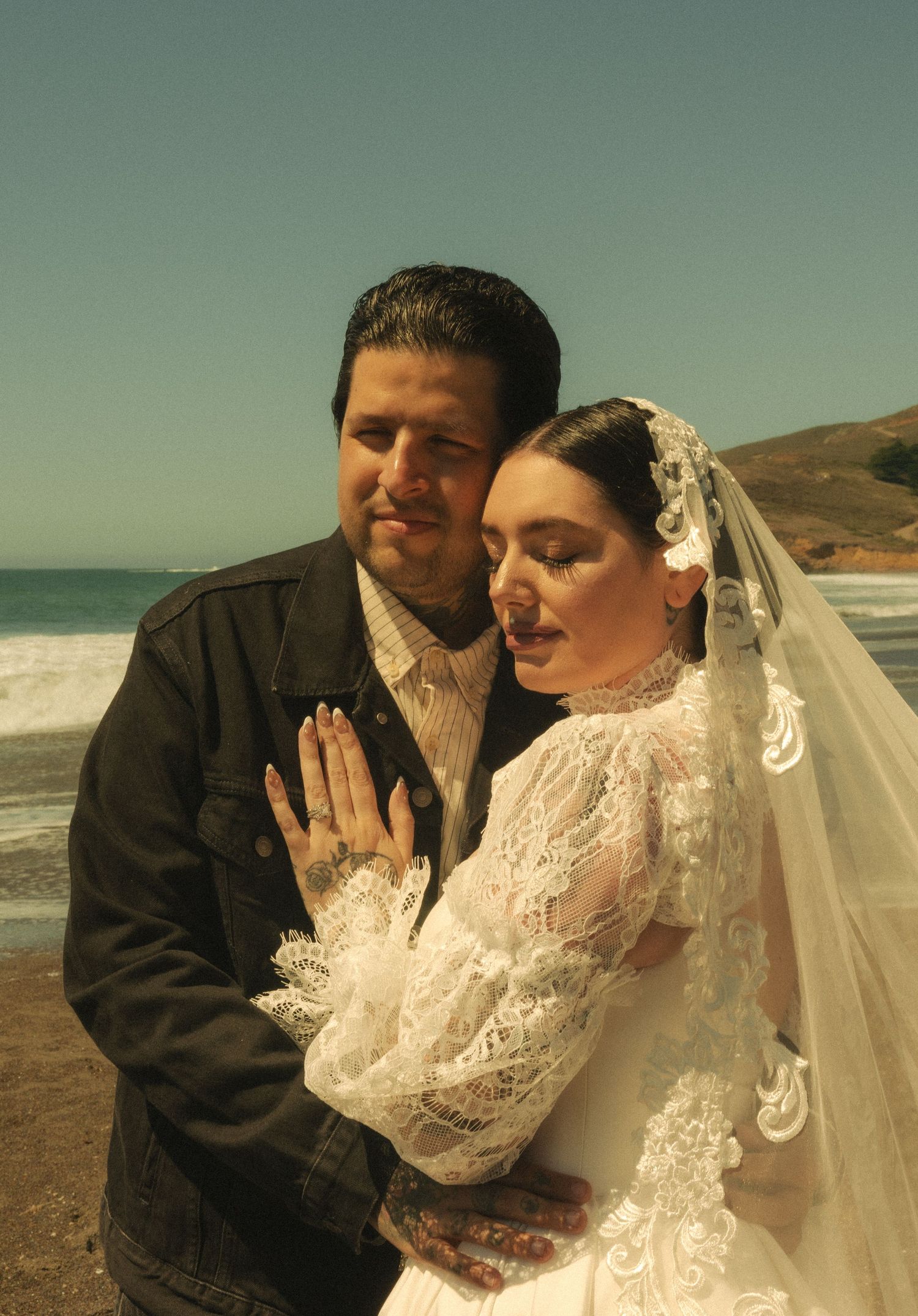 Couple in wedding attire embracing on a beach, bride in lace gown and veil, groom in dark jacket.