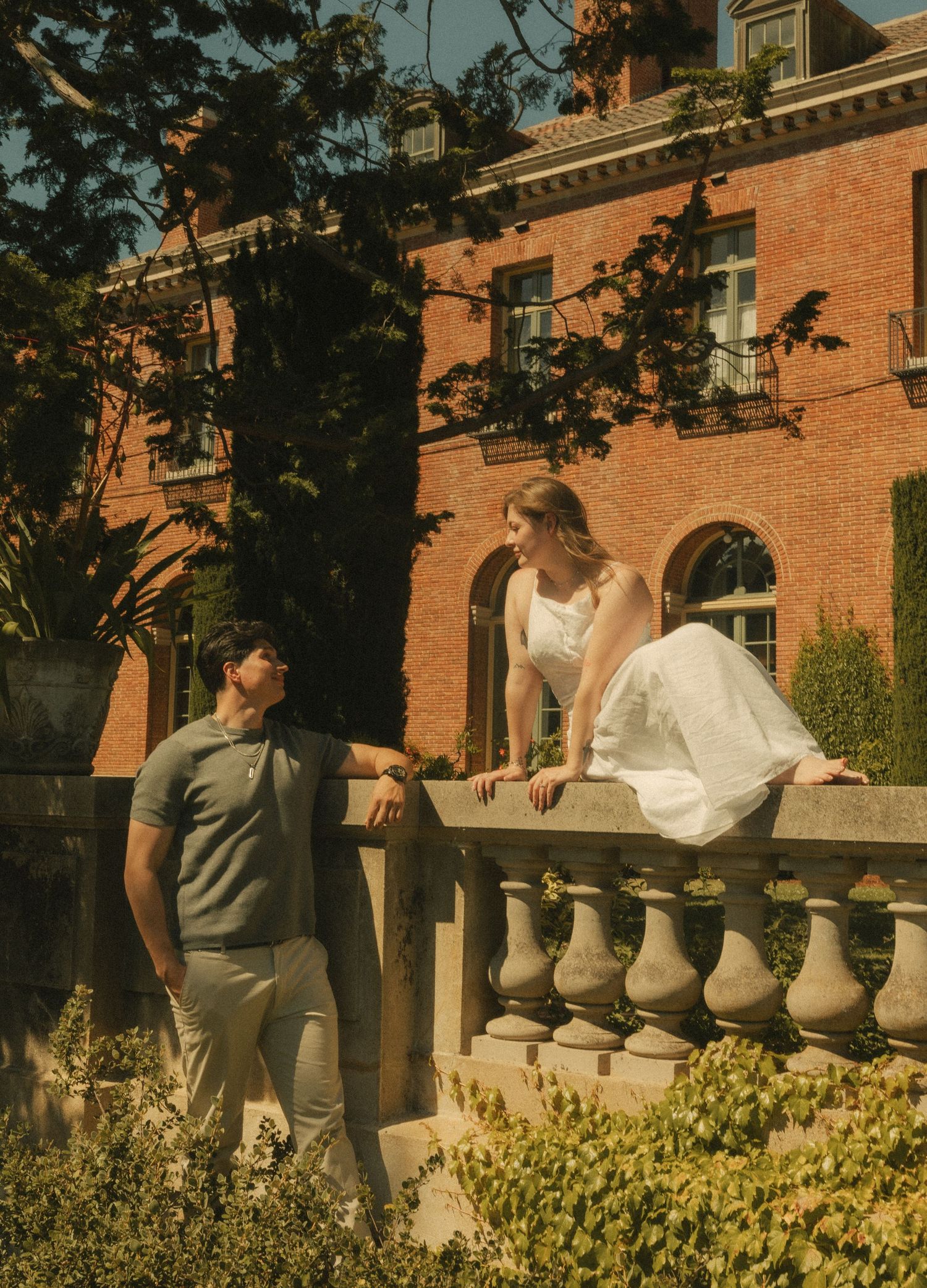 Couple posing at a brick mansion garden, woman in white dress on stone balustrade, man in polo shirt looking up at her.