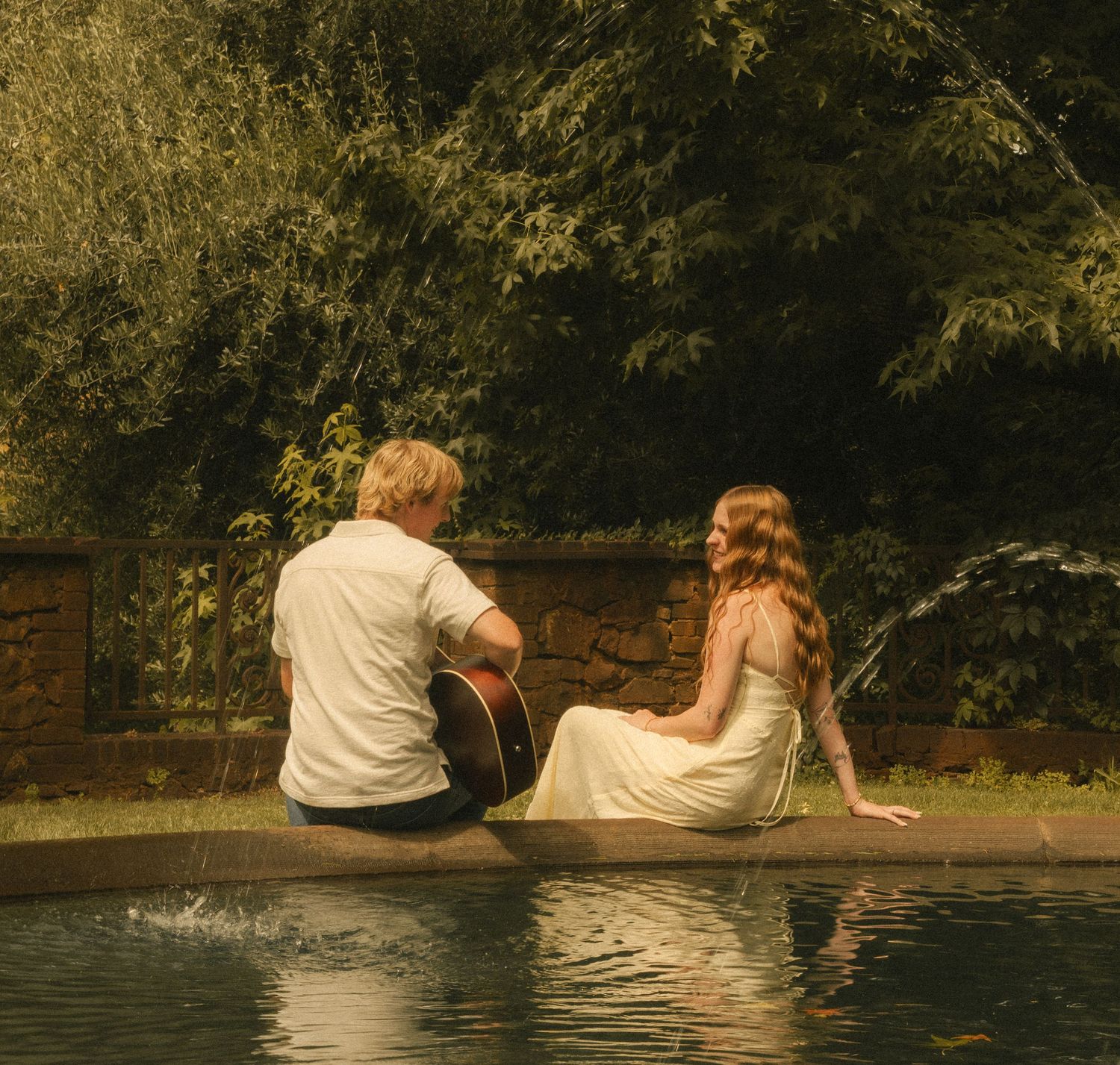 Couple sitting by a garden pool, man holding guitar, woman in white dress, surrounded by lush green trees.