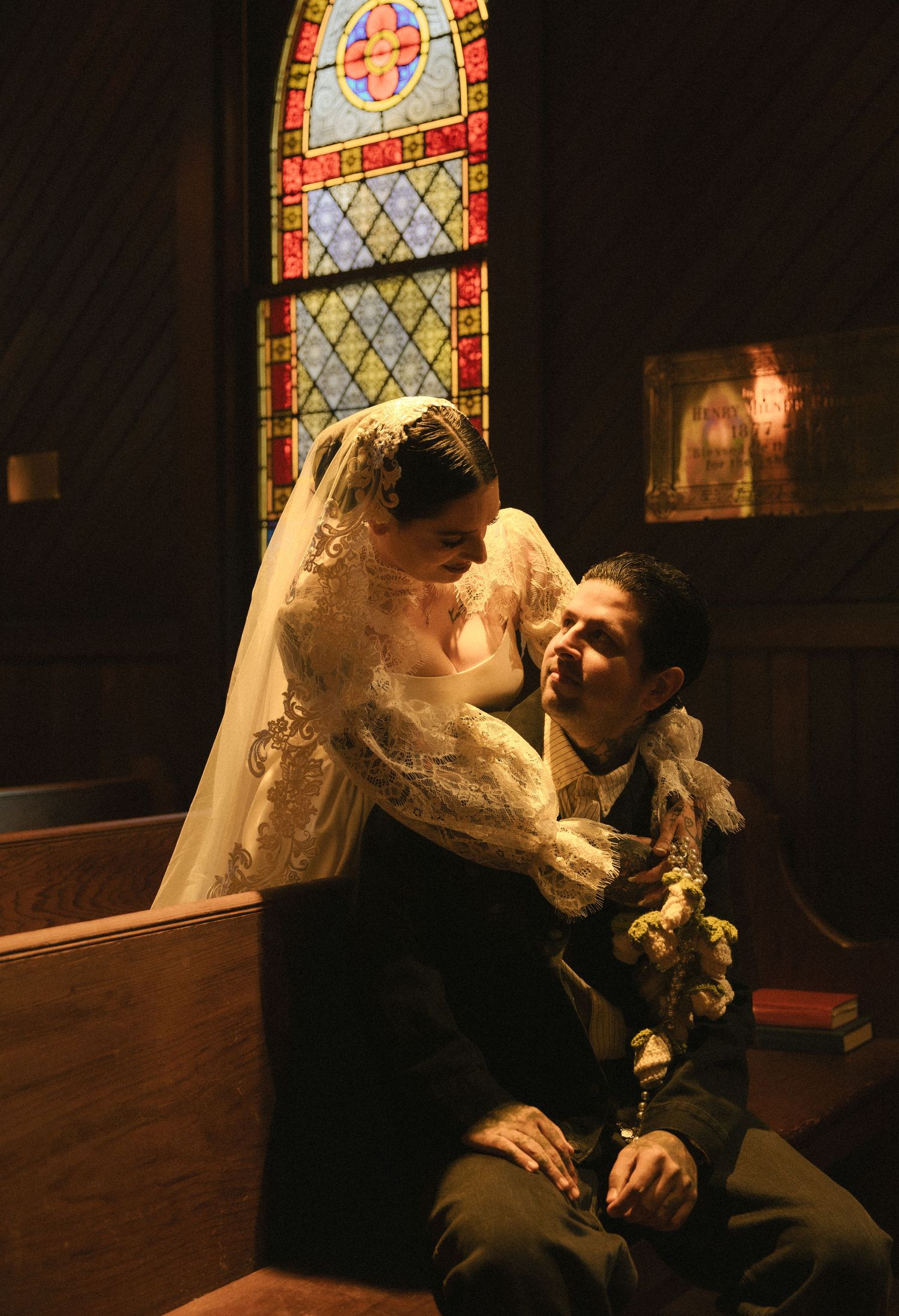 Bride and groom share a romantic moment in a church pew, bathed in warm light from colorful stained glass windows.