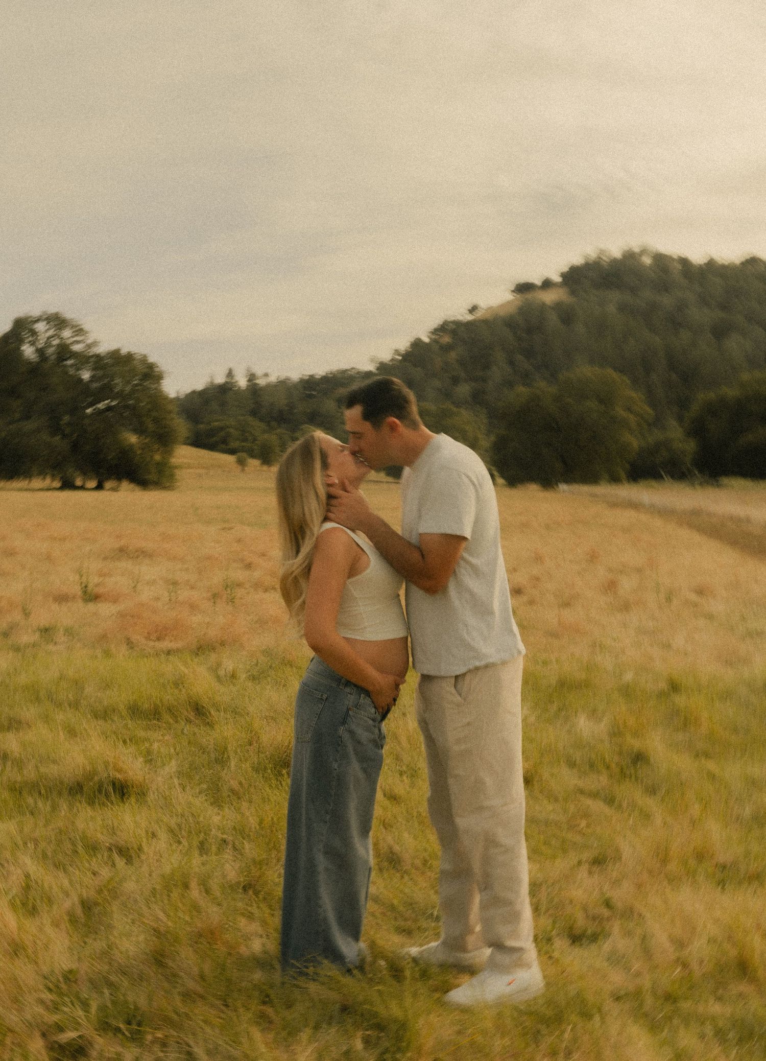 A couple shares a romantic kiss in a golden meadow with rolling hills and trees in the background.