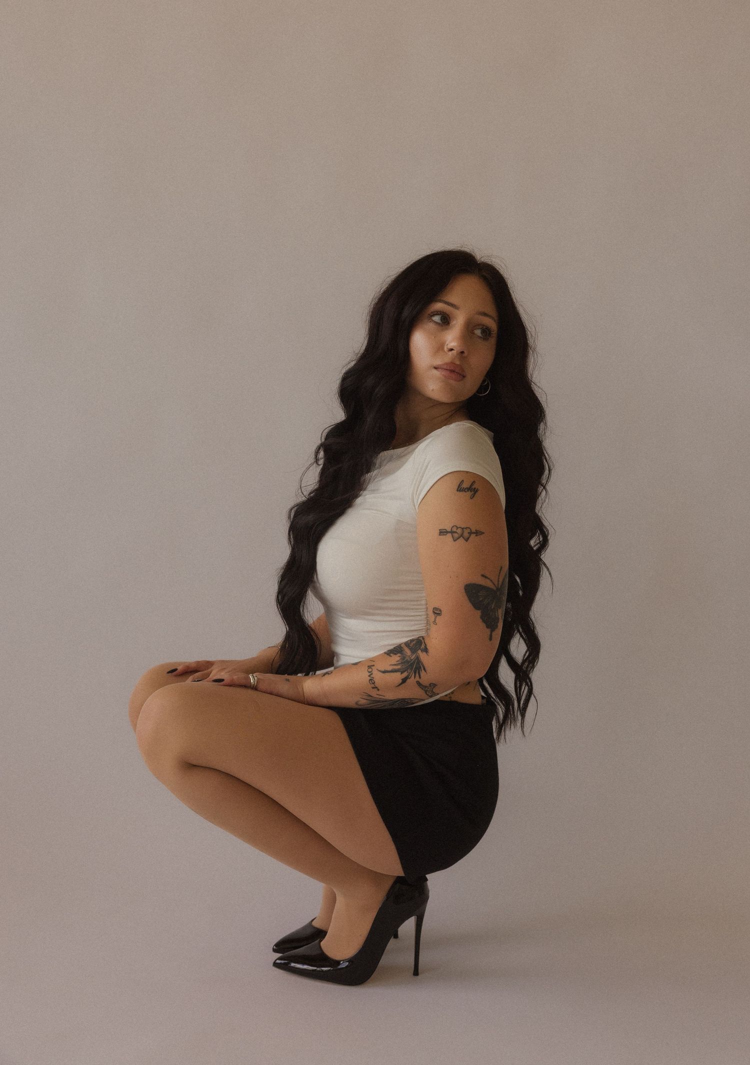 Woman with long dark hair and tattoos poses in white top, black mini skirt, and heels against neutral background.