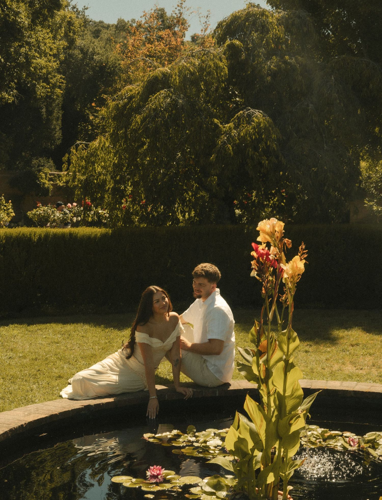 Couple in white outfits sitting by a garden pond with lily pads and colorful flowers on a sunny day.
