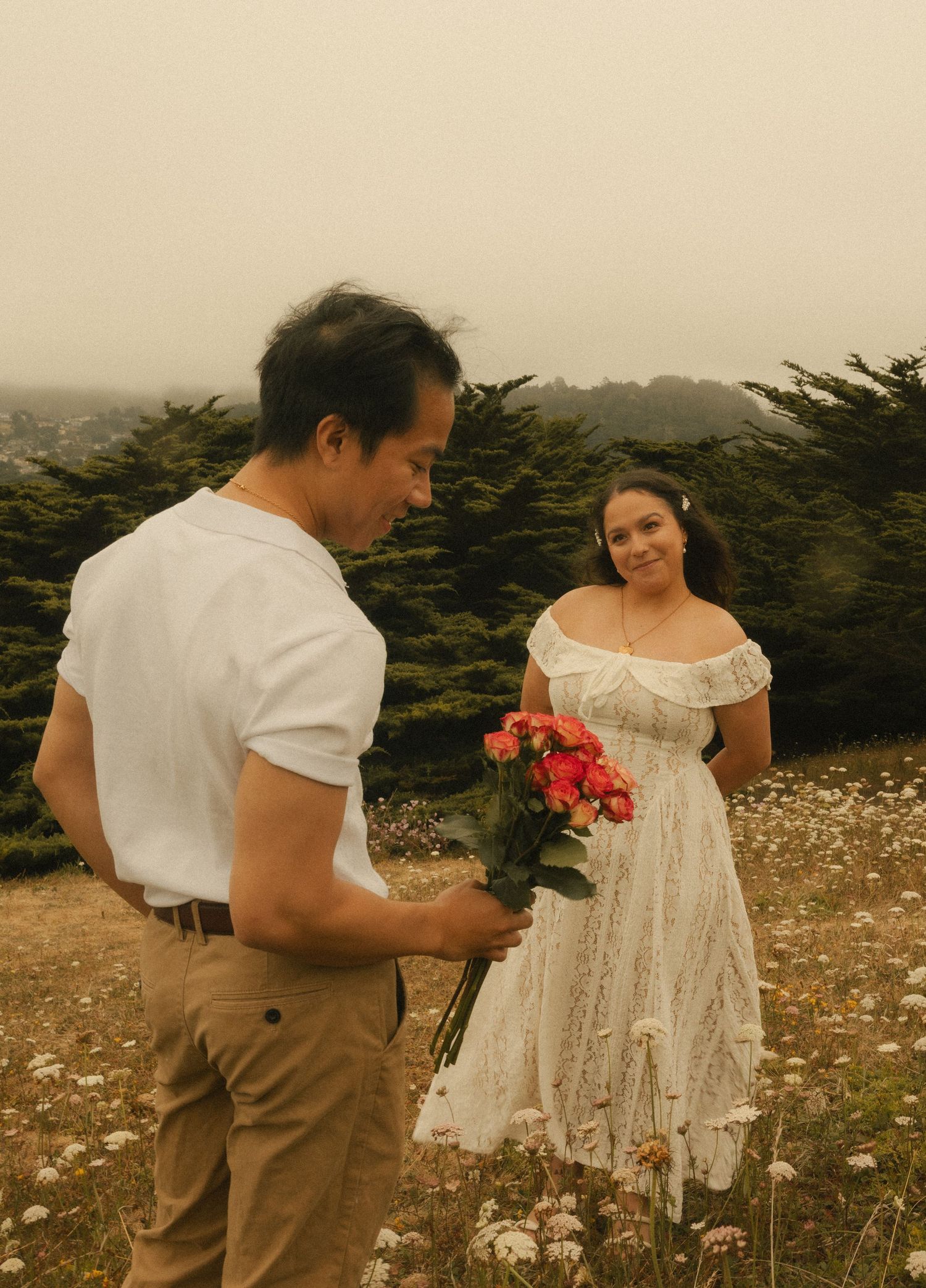 A man holds pink flowers for a smiling woman in a white dress in a foggy field with trees.