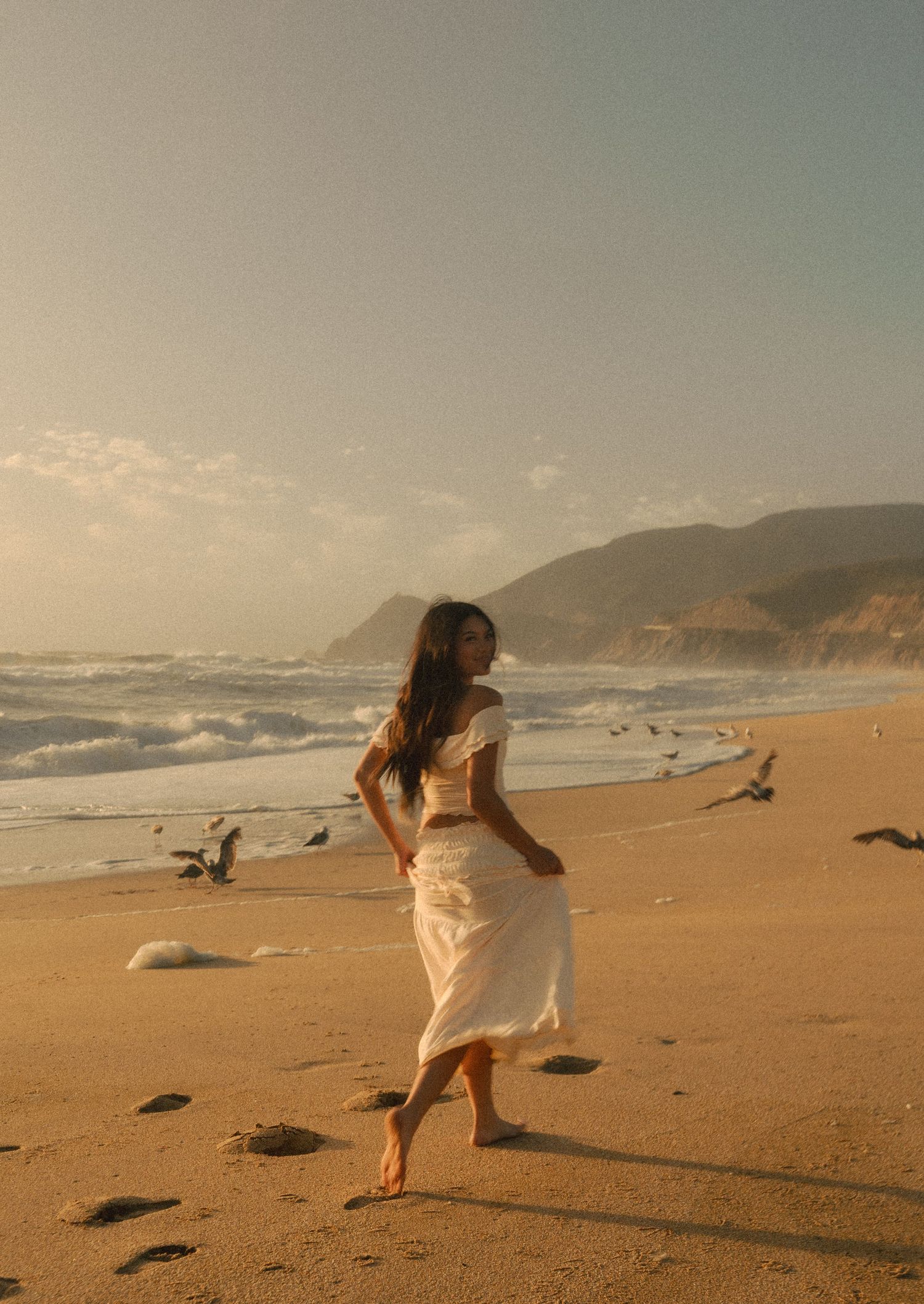 Woman in white dress walking barefoot on sandy beach at golden hour with ocean waves and cliffs in background.