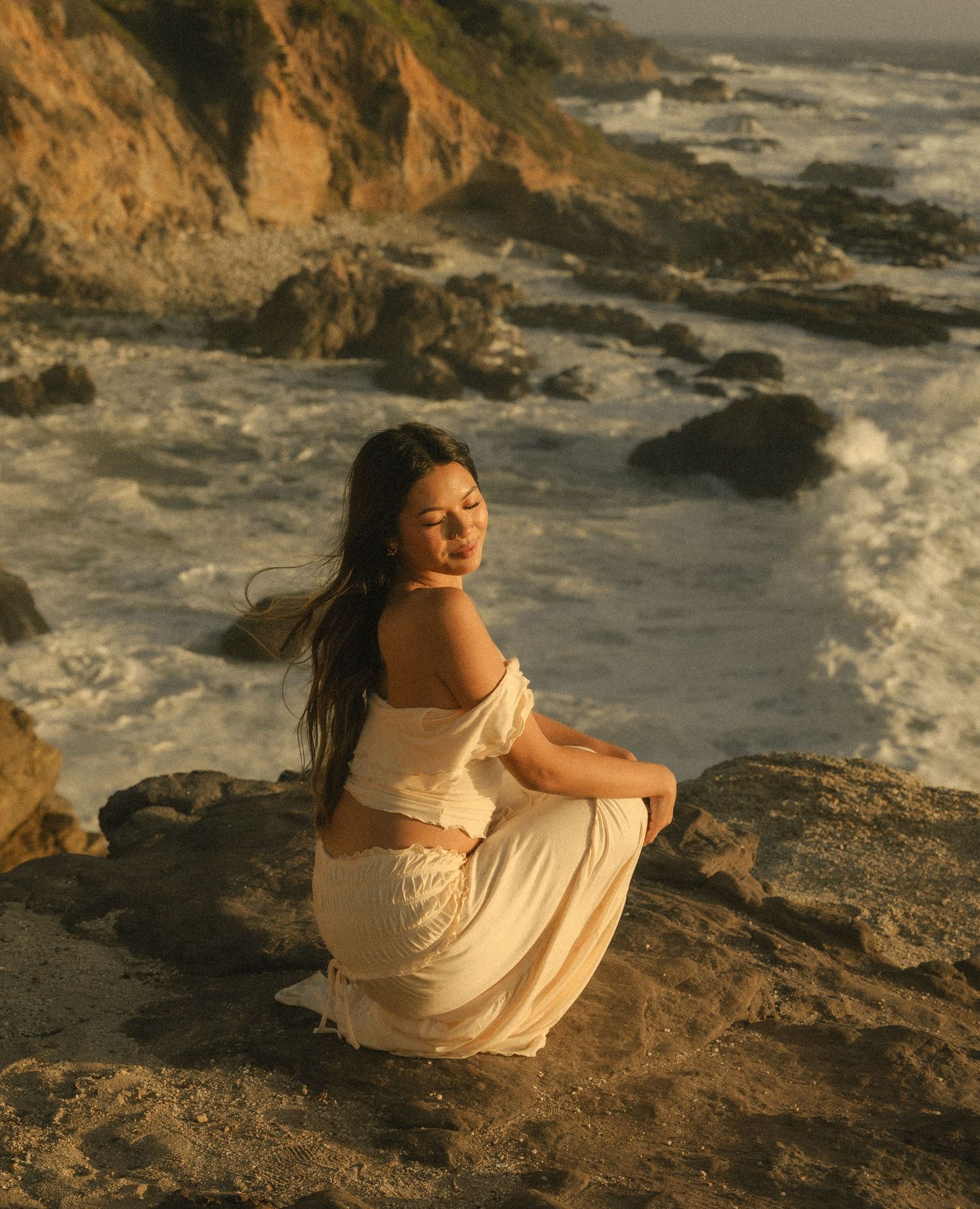 Woman in white dress sitting on coastal rocks at golden hour with ocean waves and cliffs in background.