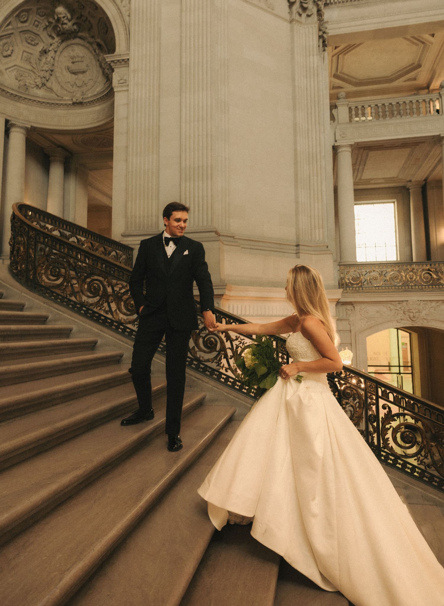 A bride in a white gown holds hands with a groom in a black tuxedo on grand marble stairs in an elegant venue.