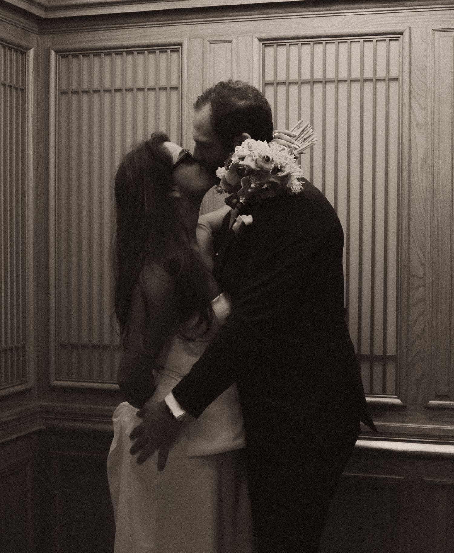 Black and white photo of a couple sharing their first kiss at a wedding ceremony, bride holding a floral bouquet.