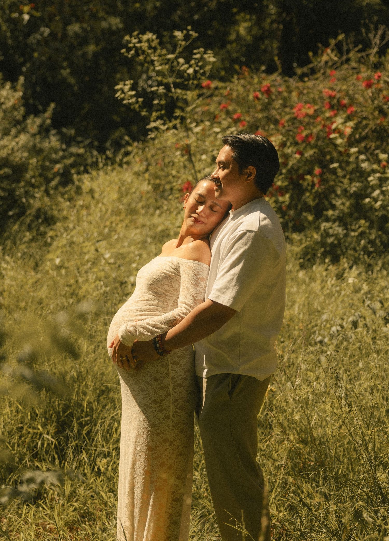 A couple embraces outdoors, the pregnant woman in a lace dress, her partner kissing her head amid lush greenery.