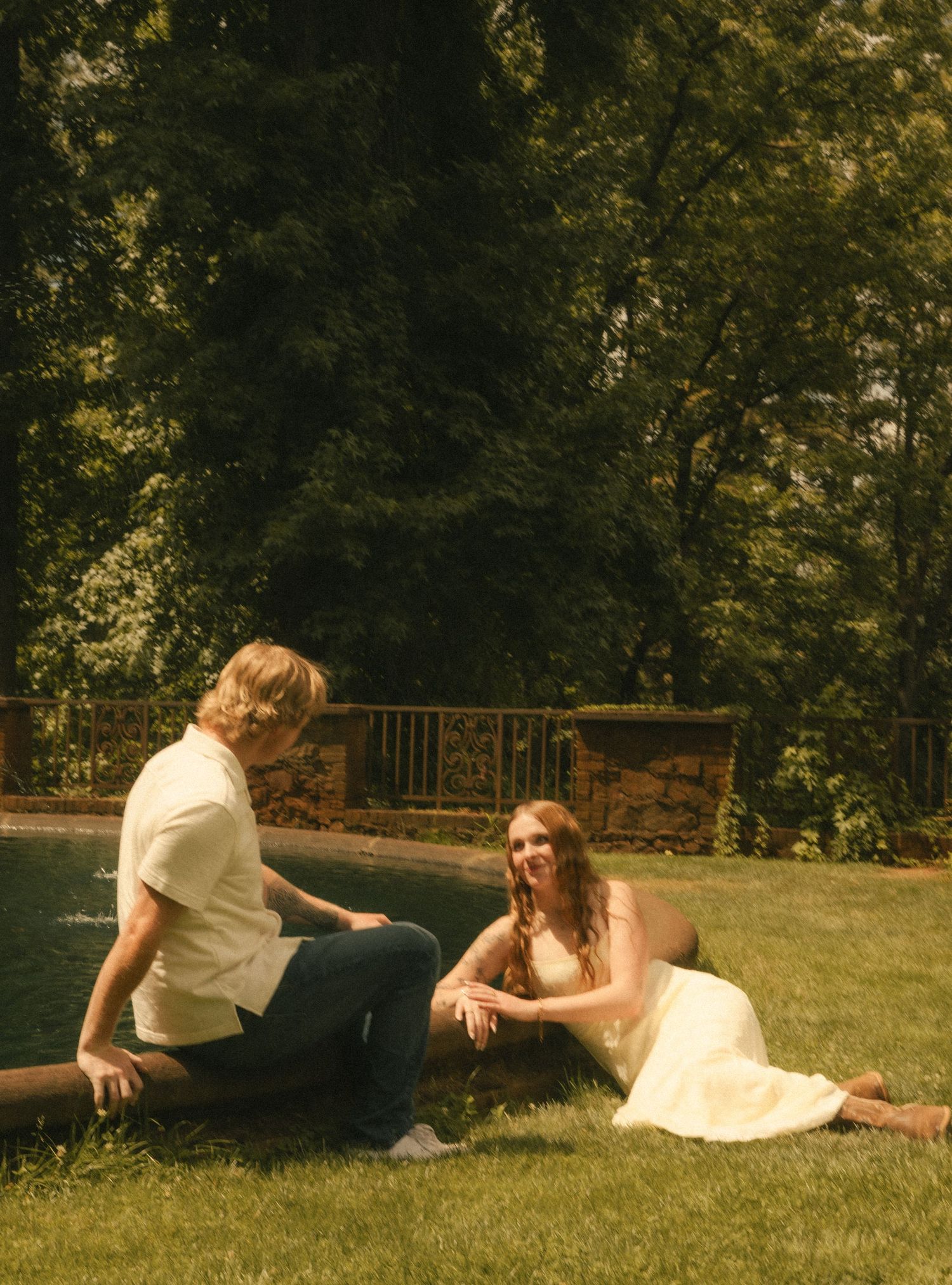 A couple sits on grass by a pool, surrounded by lush trees, sharing a joyful moment on a sunny day.