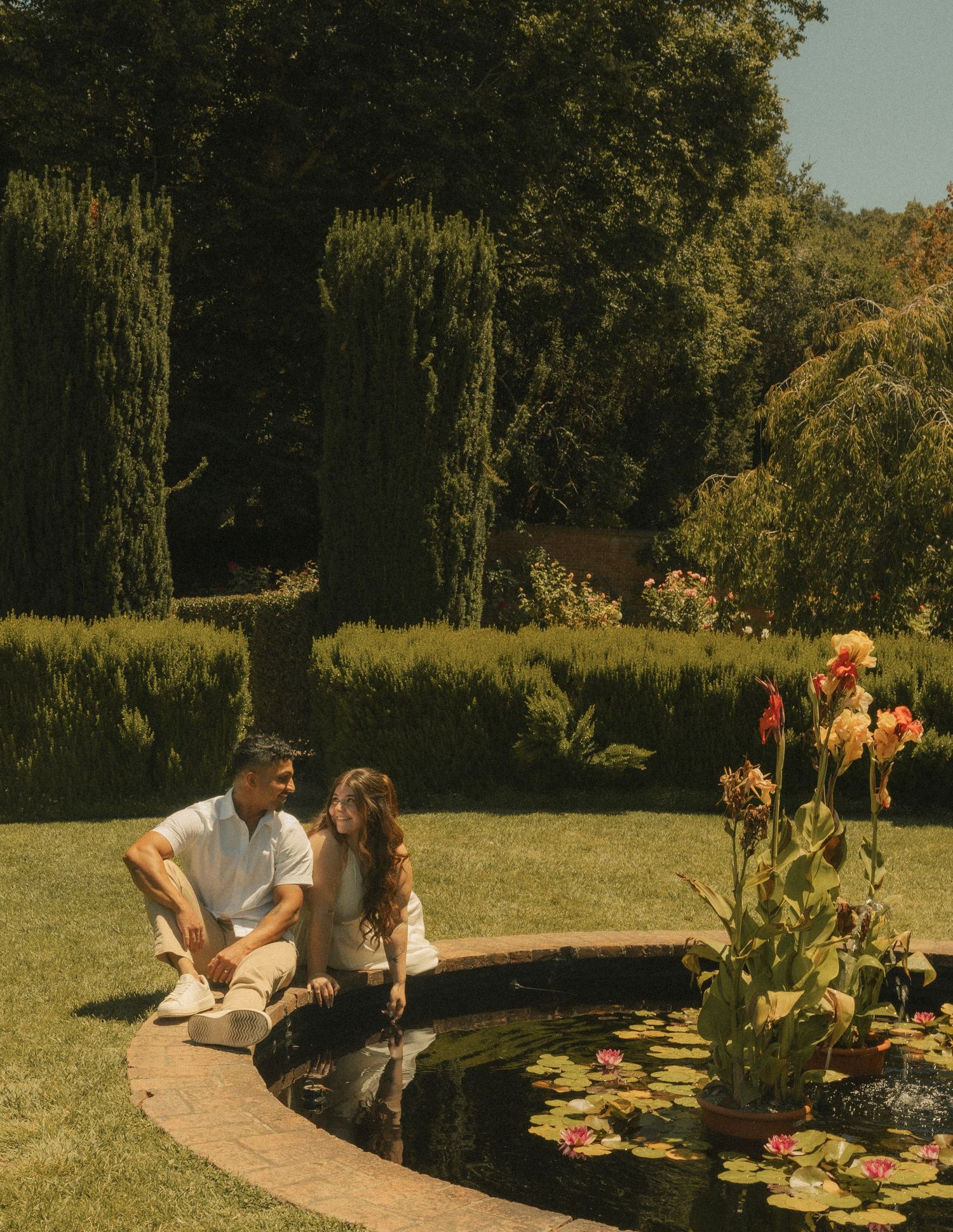 Couple sitting together by a lily pond in a lush garden with tall cypress trees and blooming flowers.