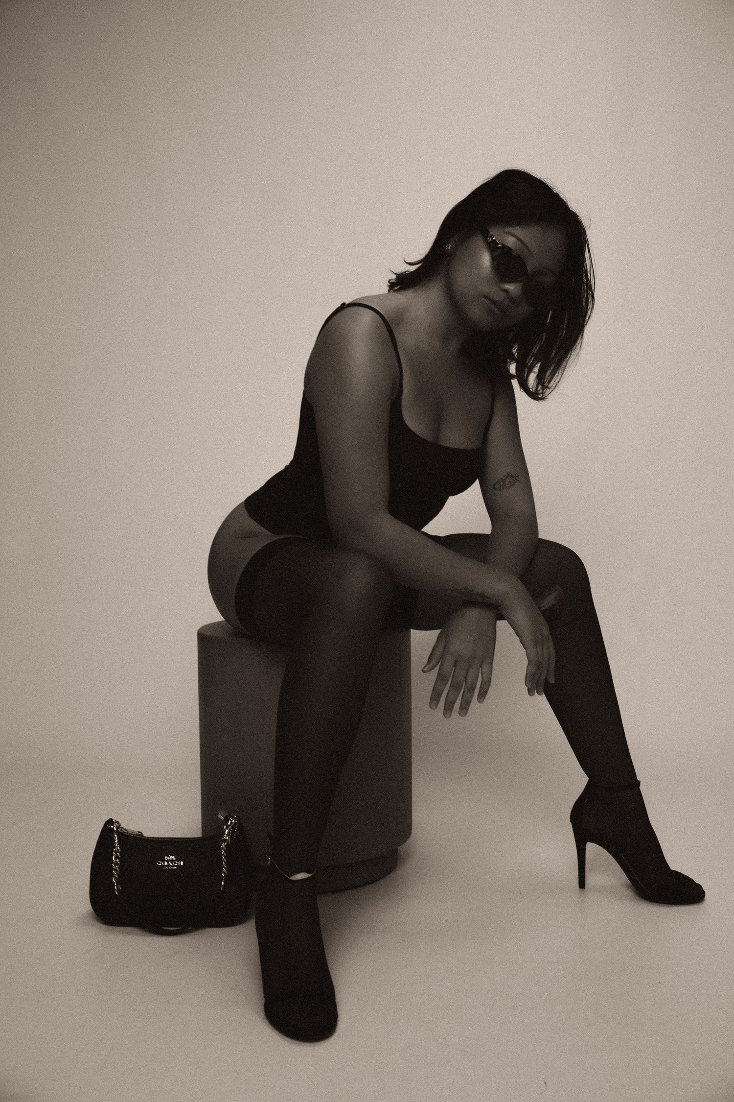 Artistic black and white portrait of person in bodysuit and heels posing on cube with designer bag on studio floor.