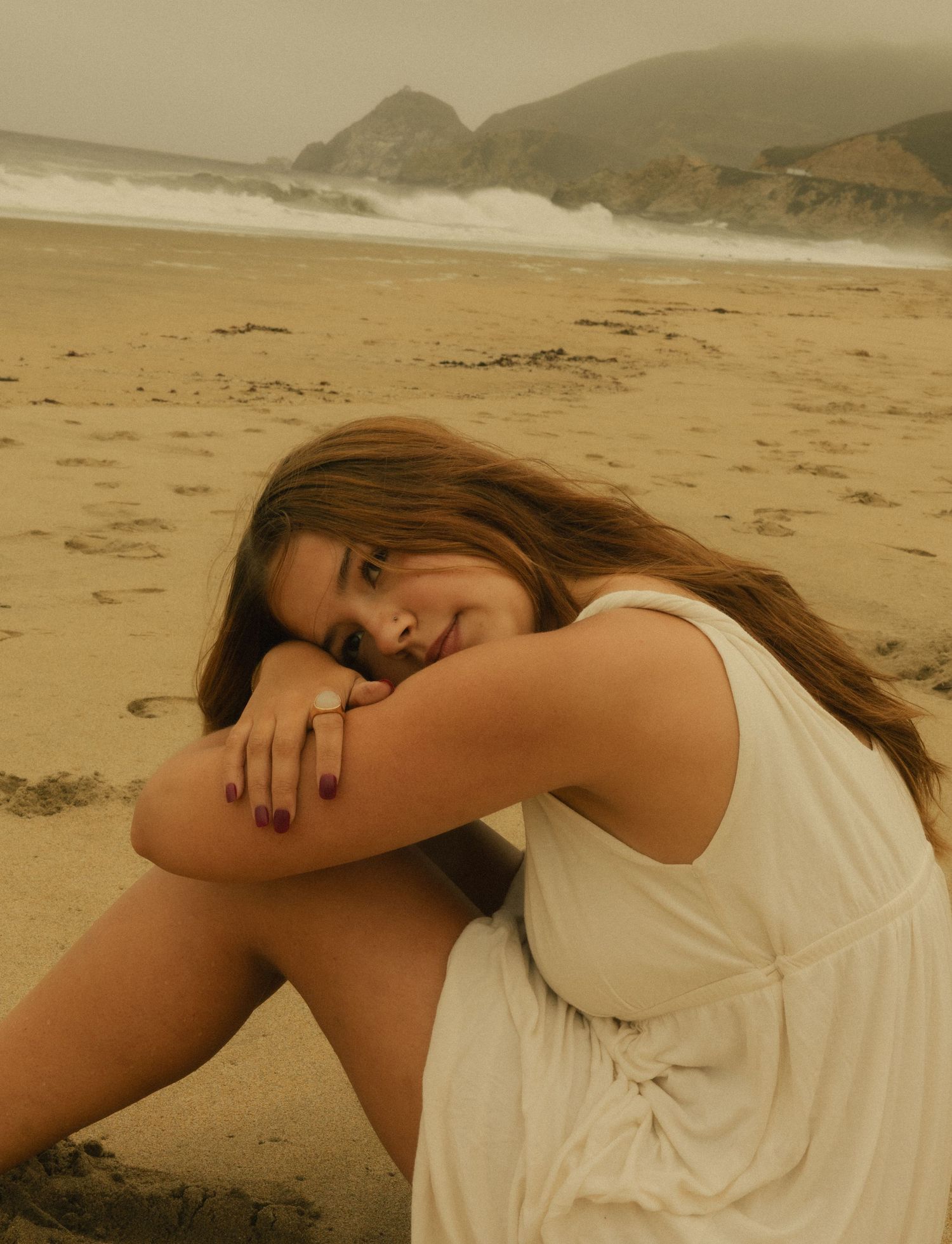 Young woman in white dress sitting on sandy beach, leaning on knees with long brown hair, ocean waves and cliffs behind her.