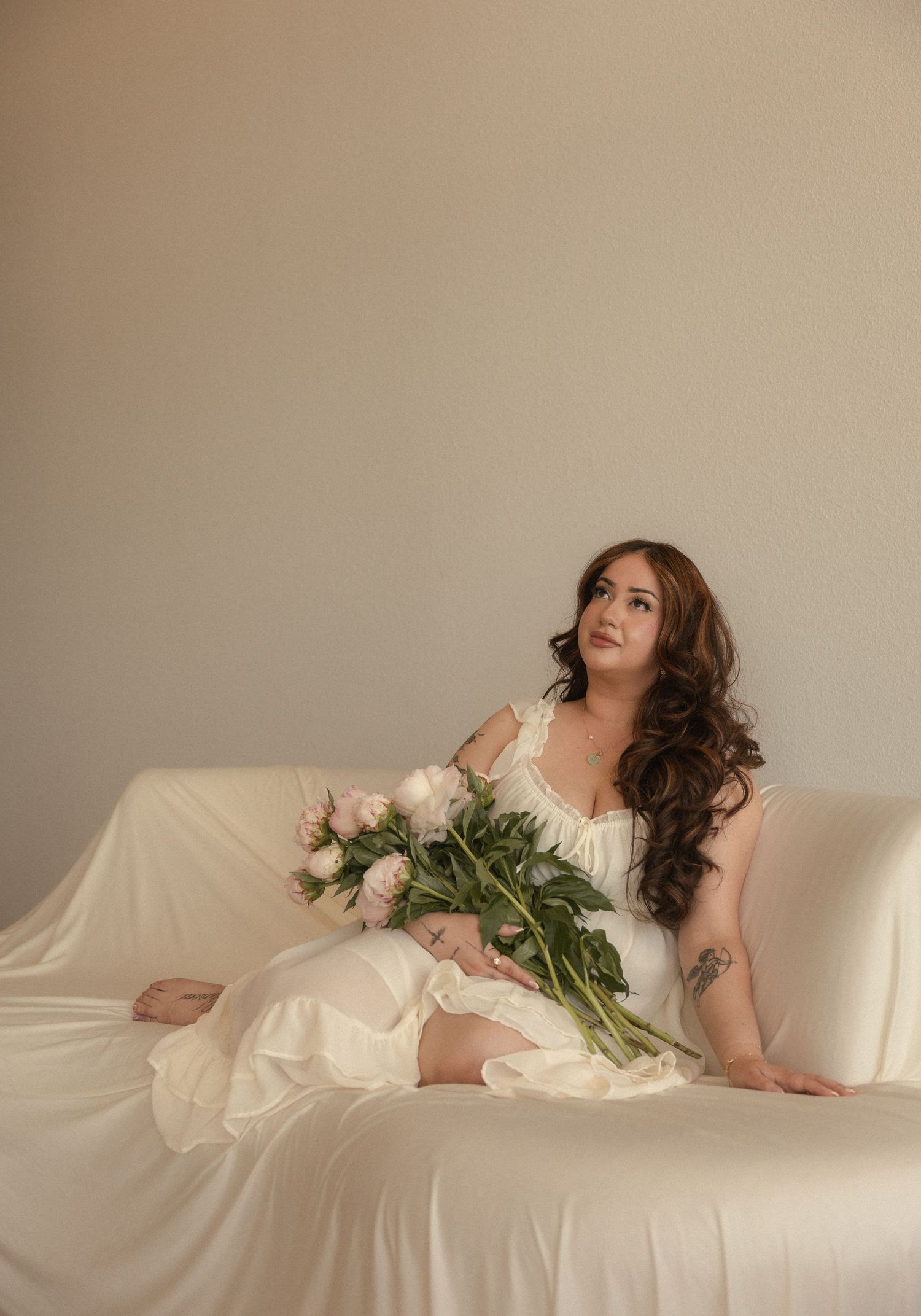 A woman in a white dress holds pink peonies while seated on a draped sofa in a soft, ethereal studio setting.