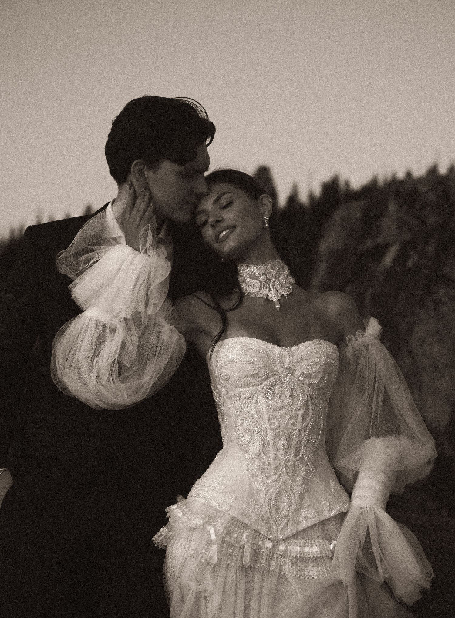 A couple in elegant wedding attire embraces outdoors, the bride in a white corset gown with tulle sleeves and lace choker.