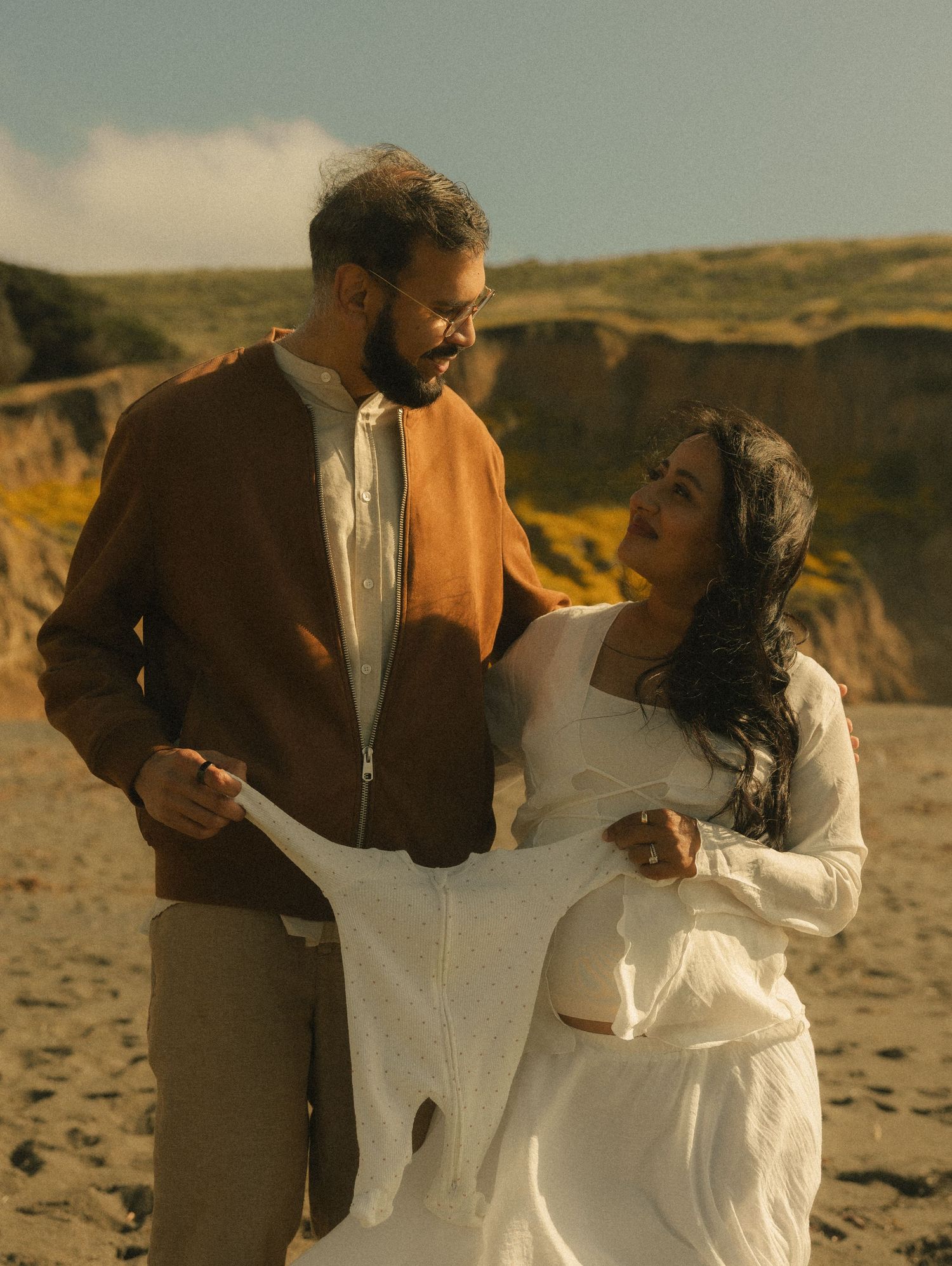 Couple sharing a tender moment on a sandy beach, woman in white dress and man in brown jacket with cliffs behind.