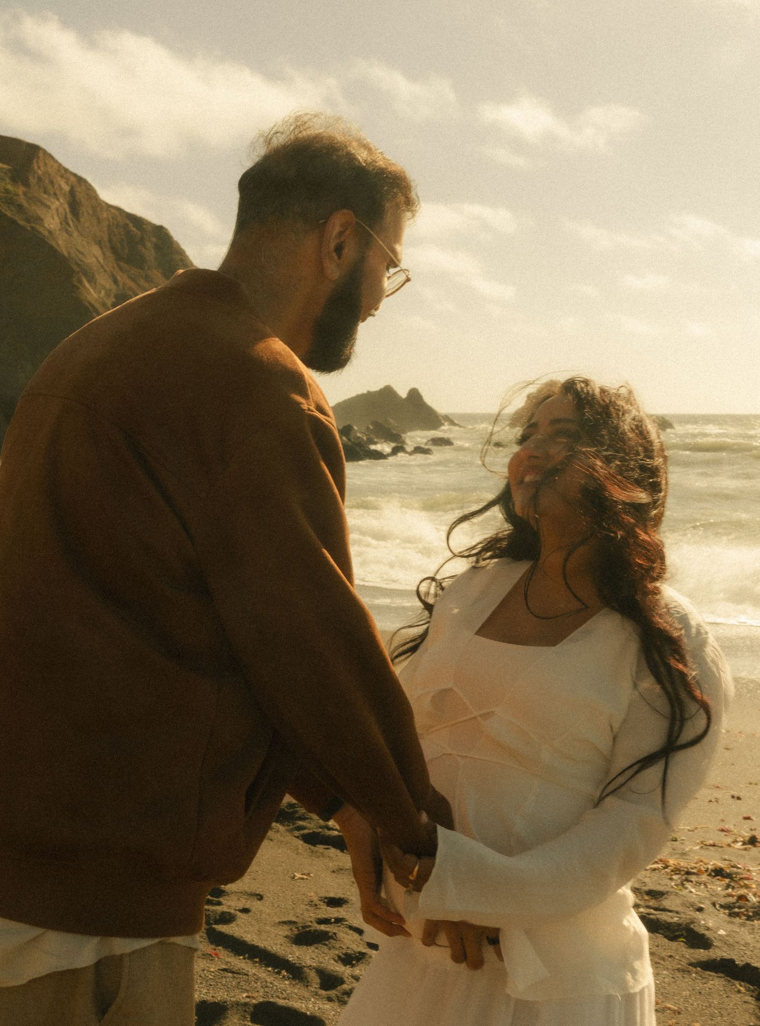 Couple holding hands on a rocky beach, man in brown jacket and woman in white dress, with ocean waves and cliffs behind them.