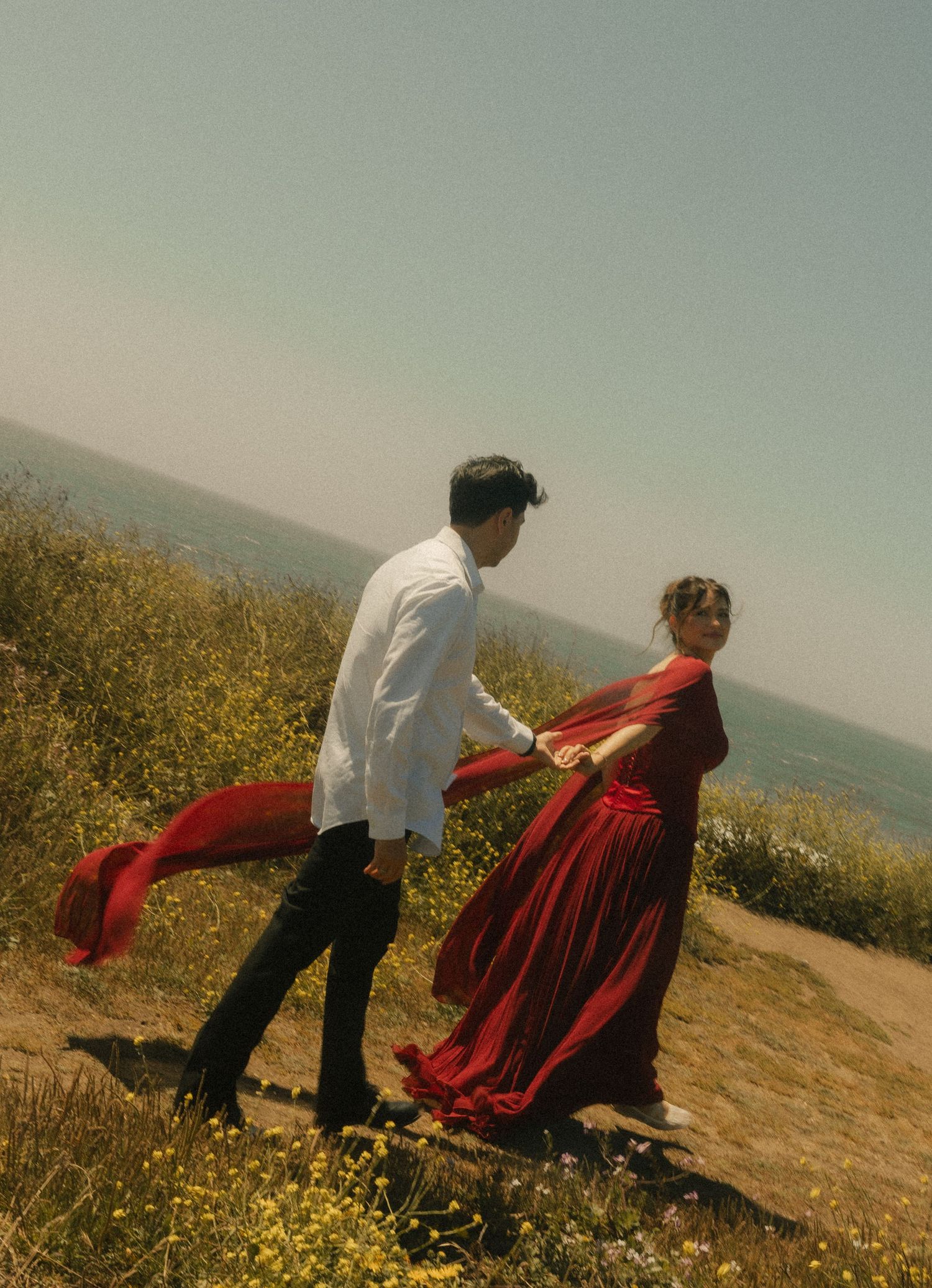 Couple in red and white outfits dancing on a hillside with wildflowers and ocean view in background.