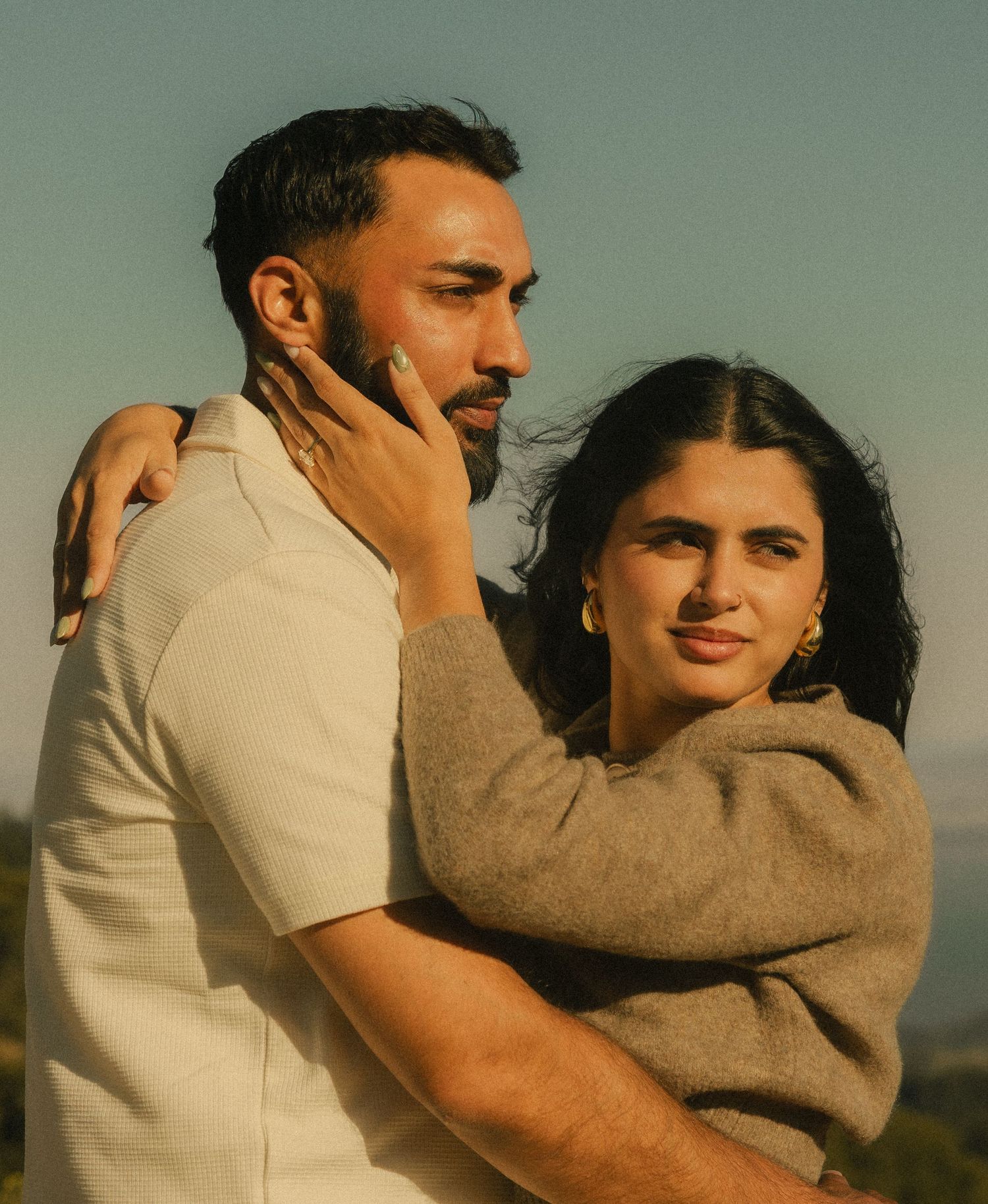A couple embraces outdoors at golden hour, woman in gray sweater with gold earrings touching mans face tenderly.