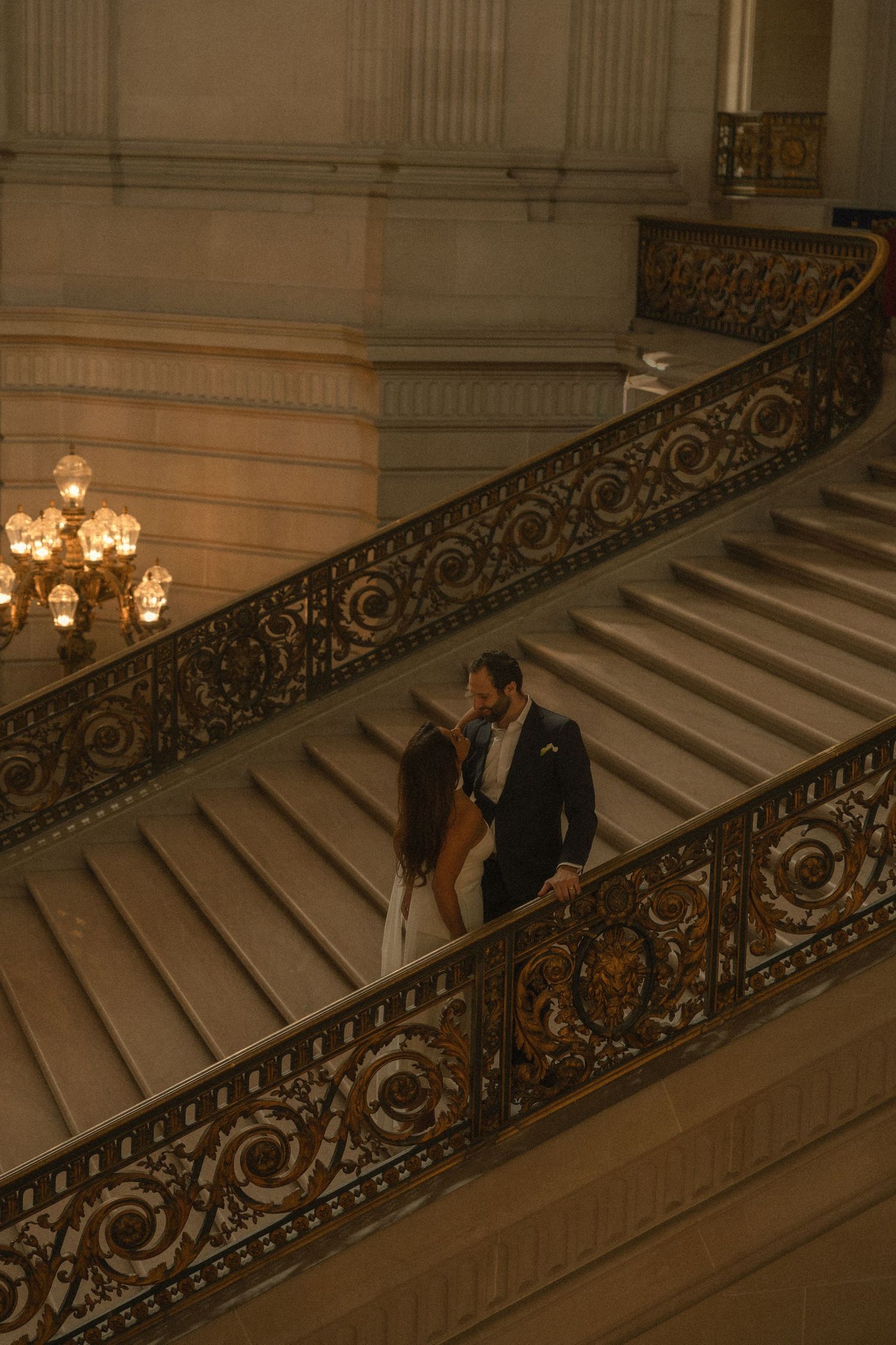 Couple sharing a romantic moment on ornate grand staircase in elegant venue, bride in white gown and groom in navy suit.