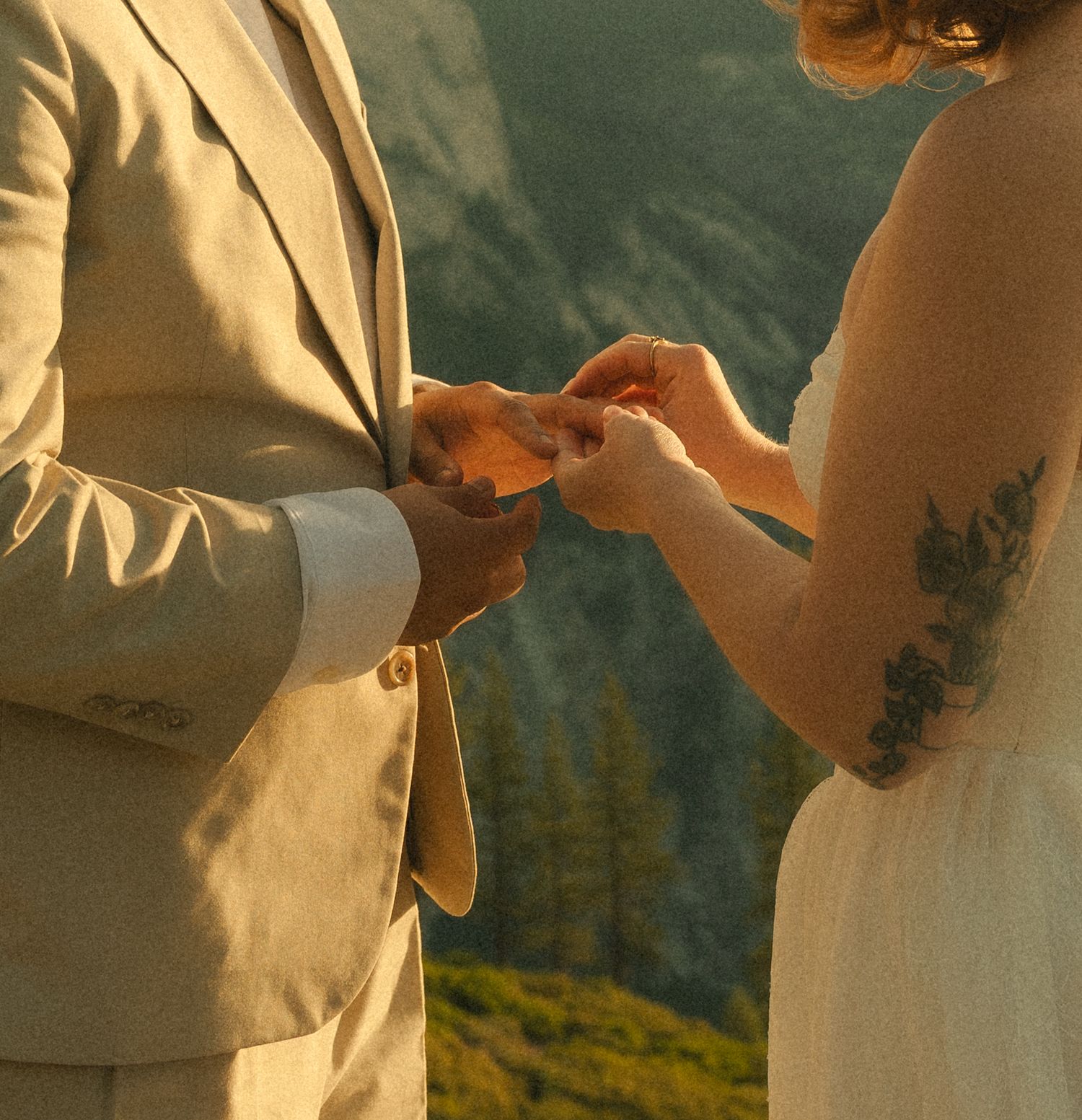 Couple exchanging rings during outdoor mountain wedding ceremony, groom in tan suit, bride with floral arm tattoo.