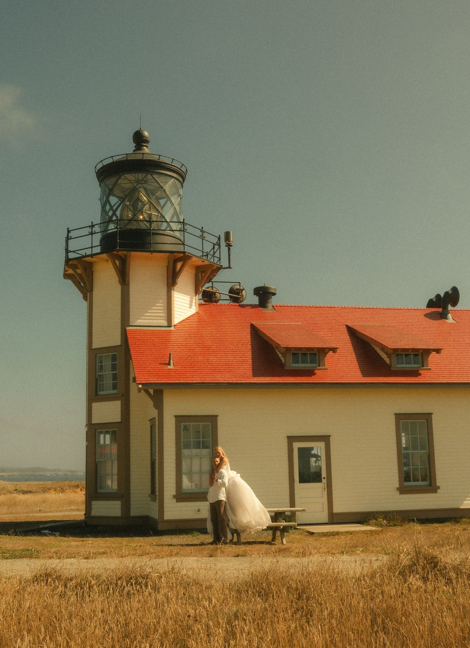 Bride in white gown poses by a red-roofed lighthouse on a sunny day with golden fields in the background.