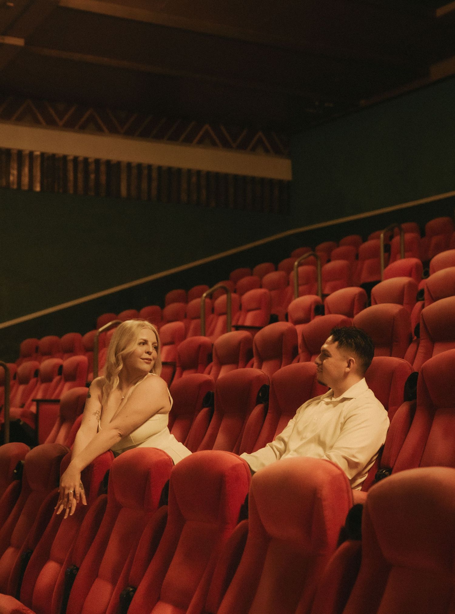 A couple sits together in red velvet theater seats in an empty vintage cinema, sharing a romantic moment.