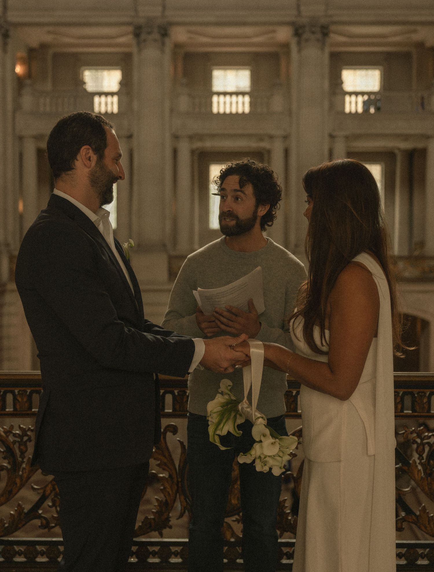 Couple exchanging vows at elegant indoor wedding ceremony with officiant holding papers in grand architectural venue.