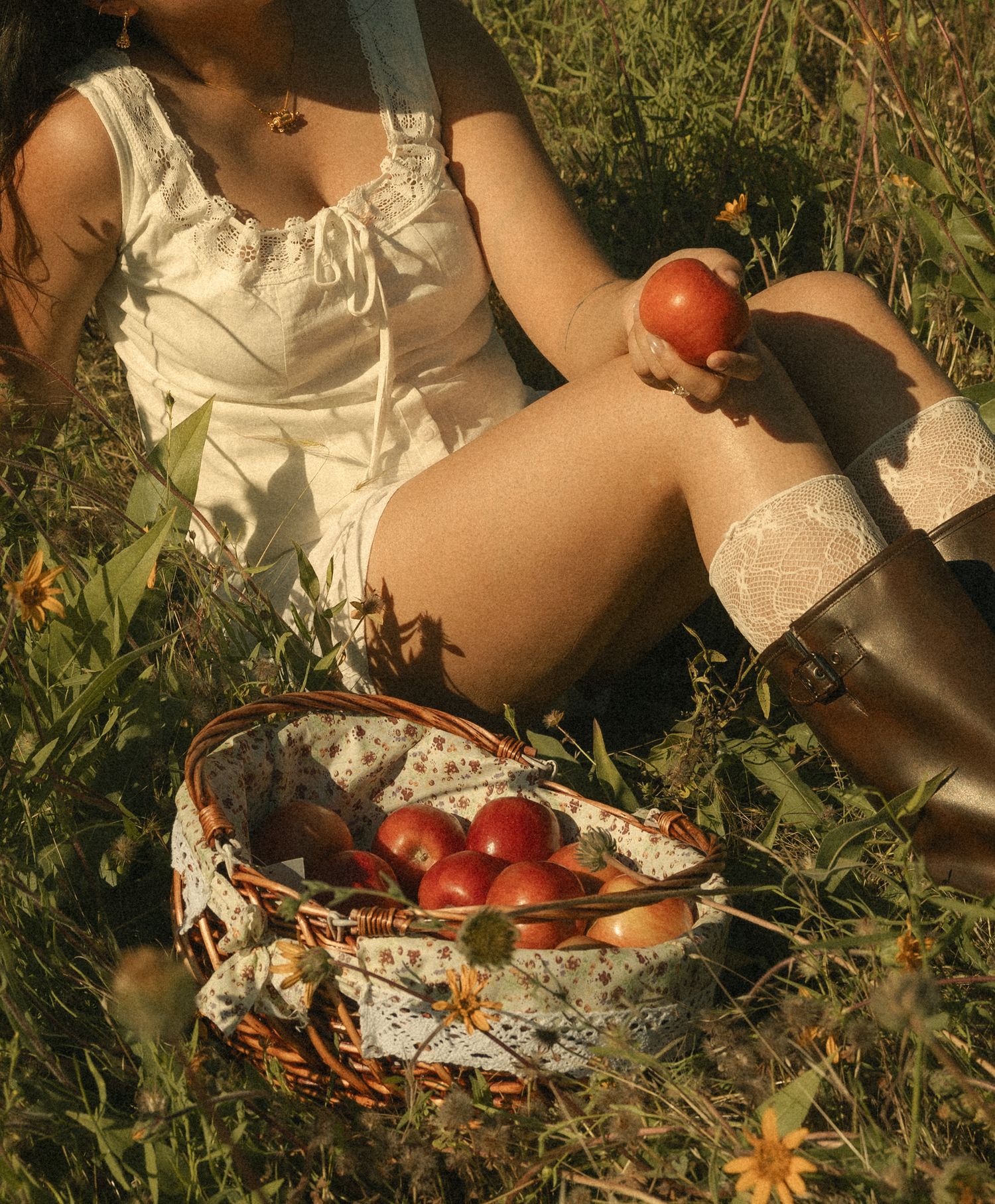 Woman in white dress sitting in grass holding a red apple beside a wicker basket full of fresh apples.
