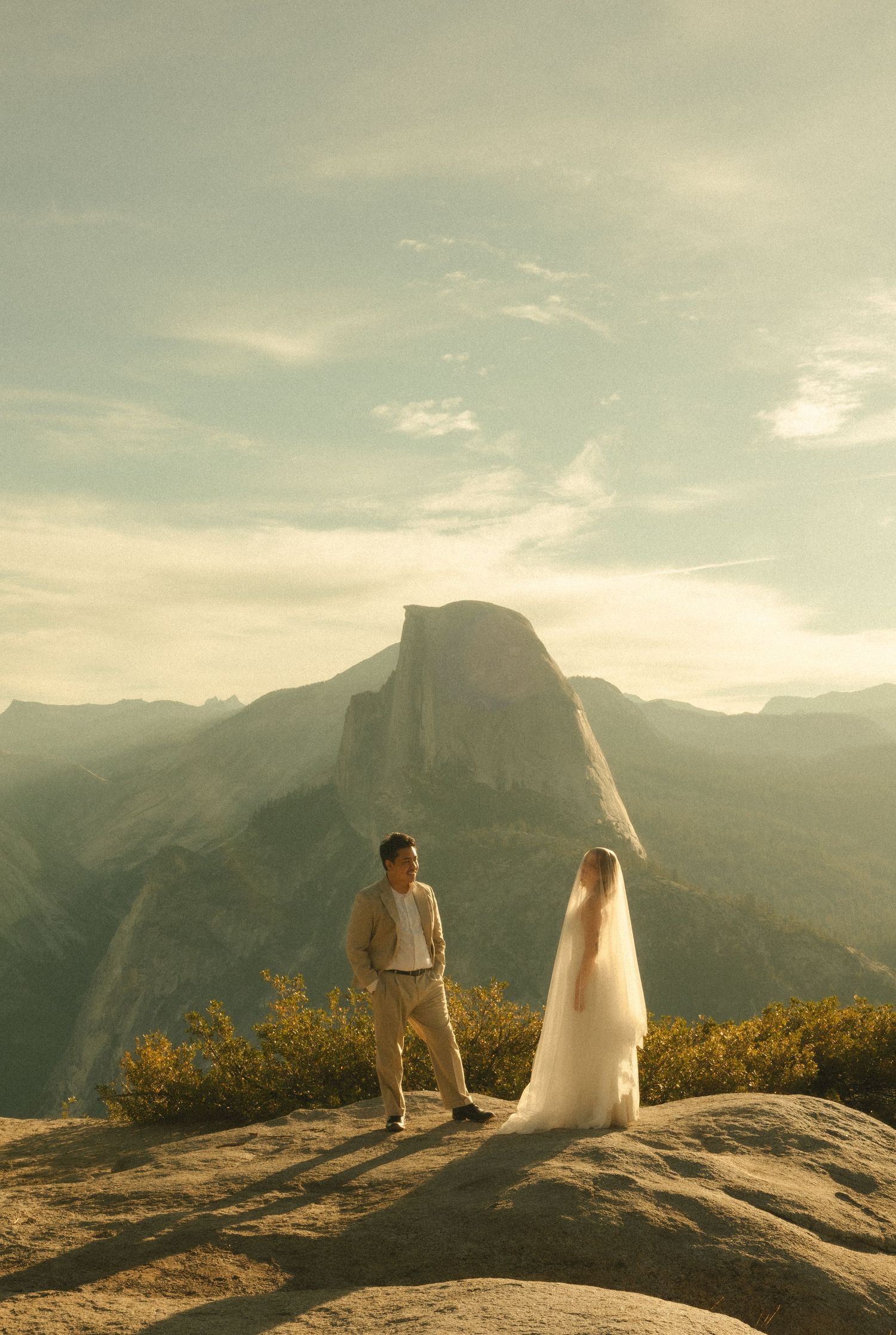 Couple in wedding attire standing on rocky overlook with Half Dome and Yosemite Valley at golden hour.