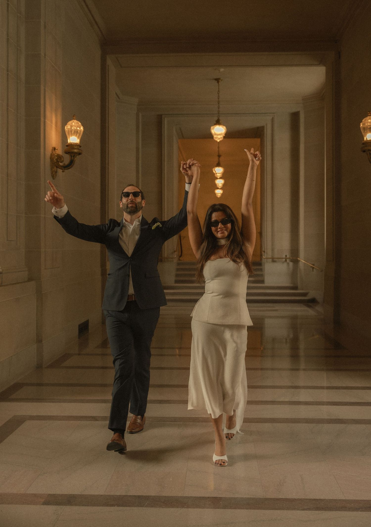 Joyful newlyweds celebrate in elegant hall, bride in white dress and groom in dark suit, arms raised in triumph.
