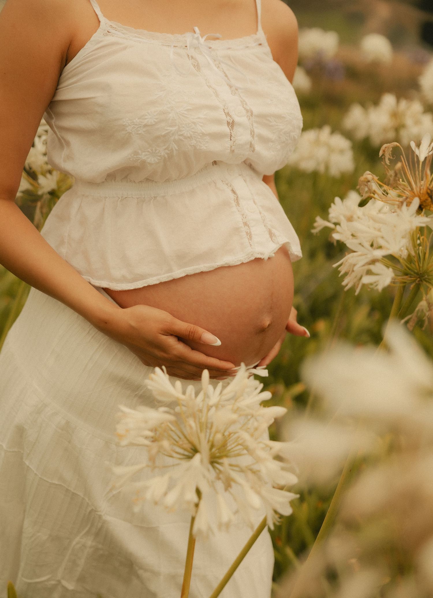 Pregnant woman in white crop top and skirt cradling baby bump among white flowers in a serene garden.
