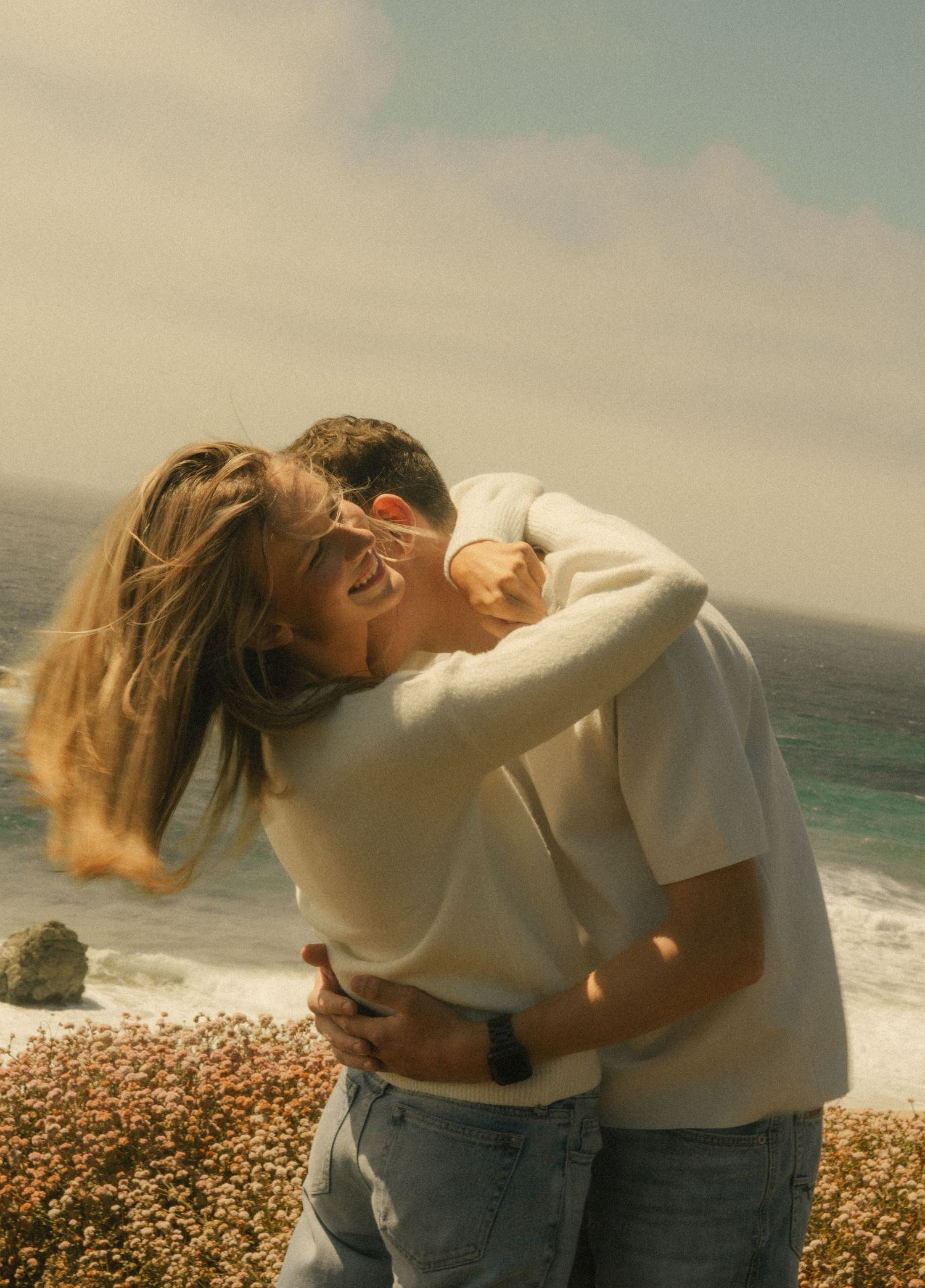 Couple embracing and laughing on a windy coastal cliffside with ocean waves and wildflowers in the background.