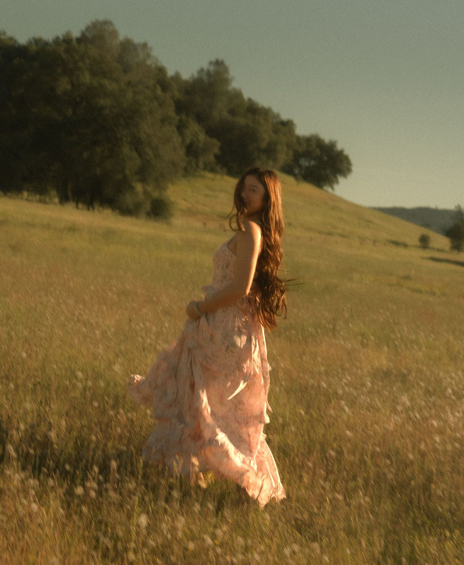 A woman in a flowing pink floral dress walks through a sunlit golden meadow with green hills and trees in the background.