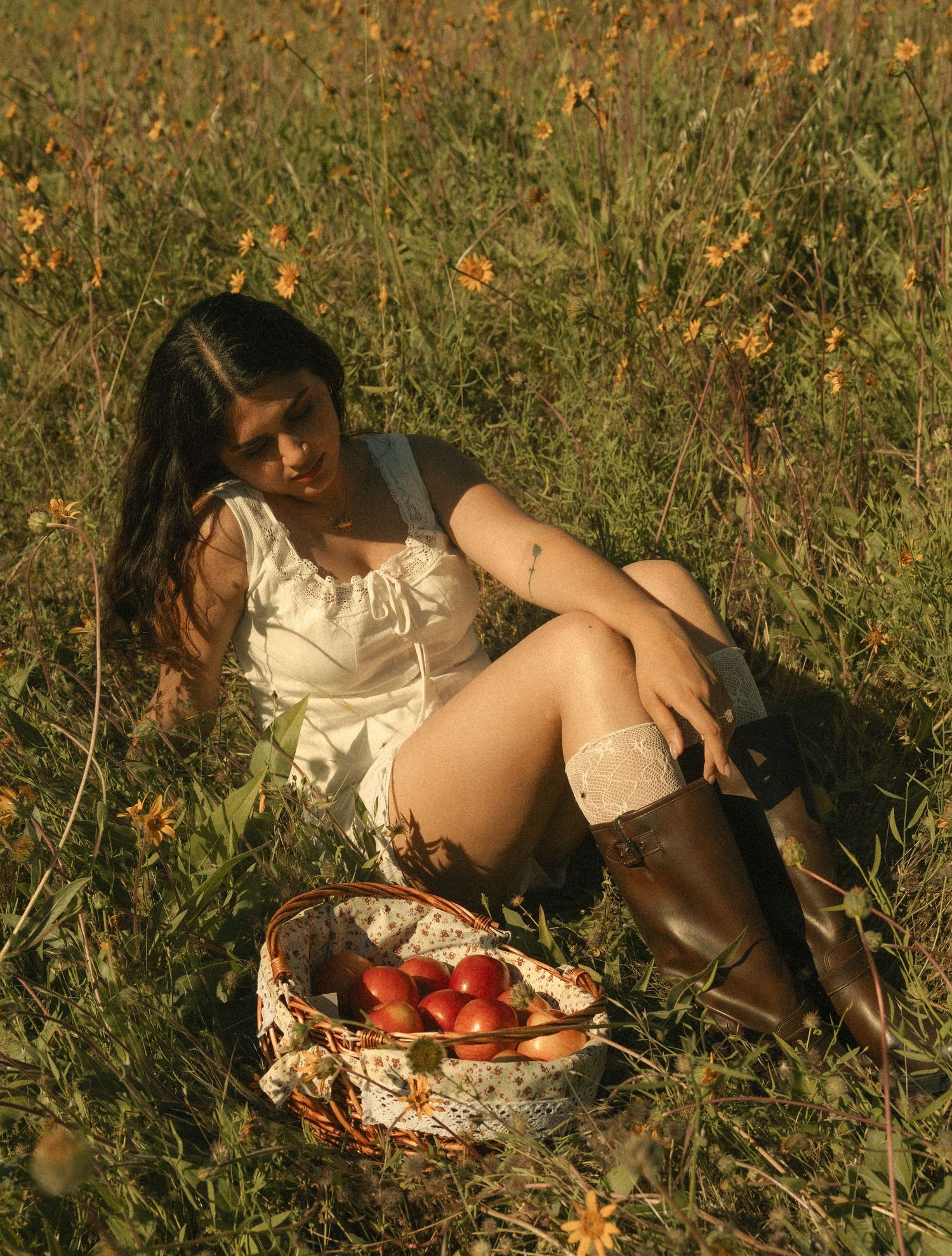 Young woman in white dress and boots sits in a wildflower field with a basket of fresh red apples.