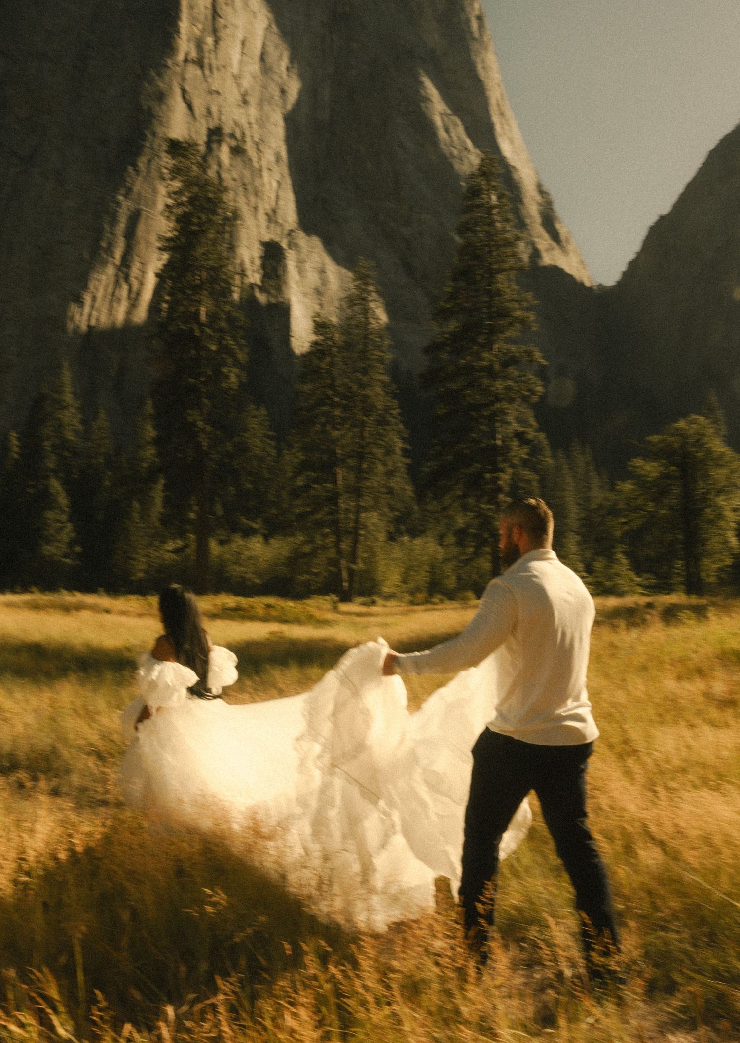 Couple dancing in wedding attire in a golden meadow with dramatic granite cliffs of Yosemite Valley behind them.
