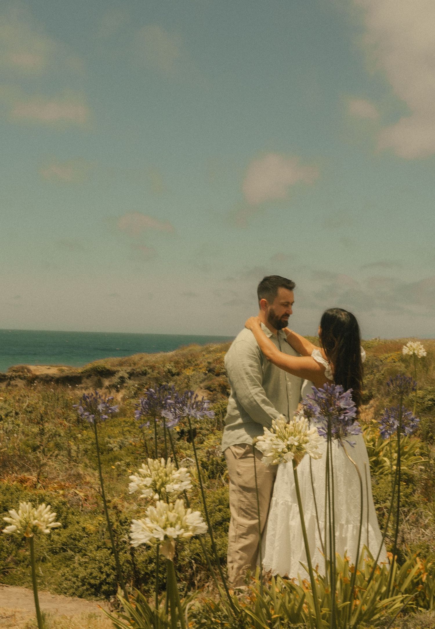 Couple embracing among white and purple agapanthus flowers with ocean views on a sunny coastal day.