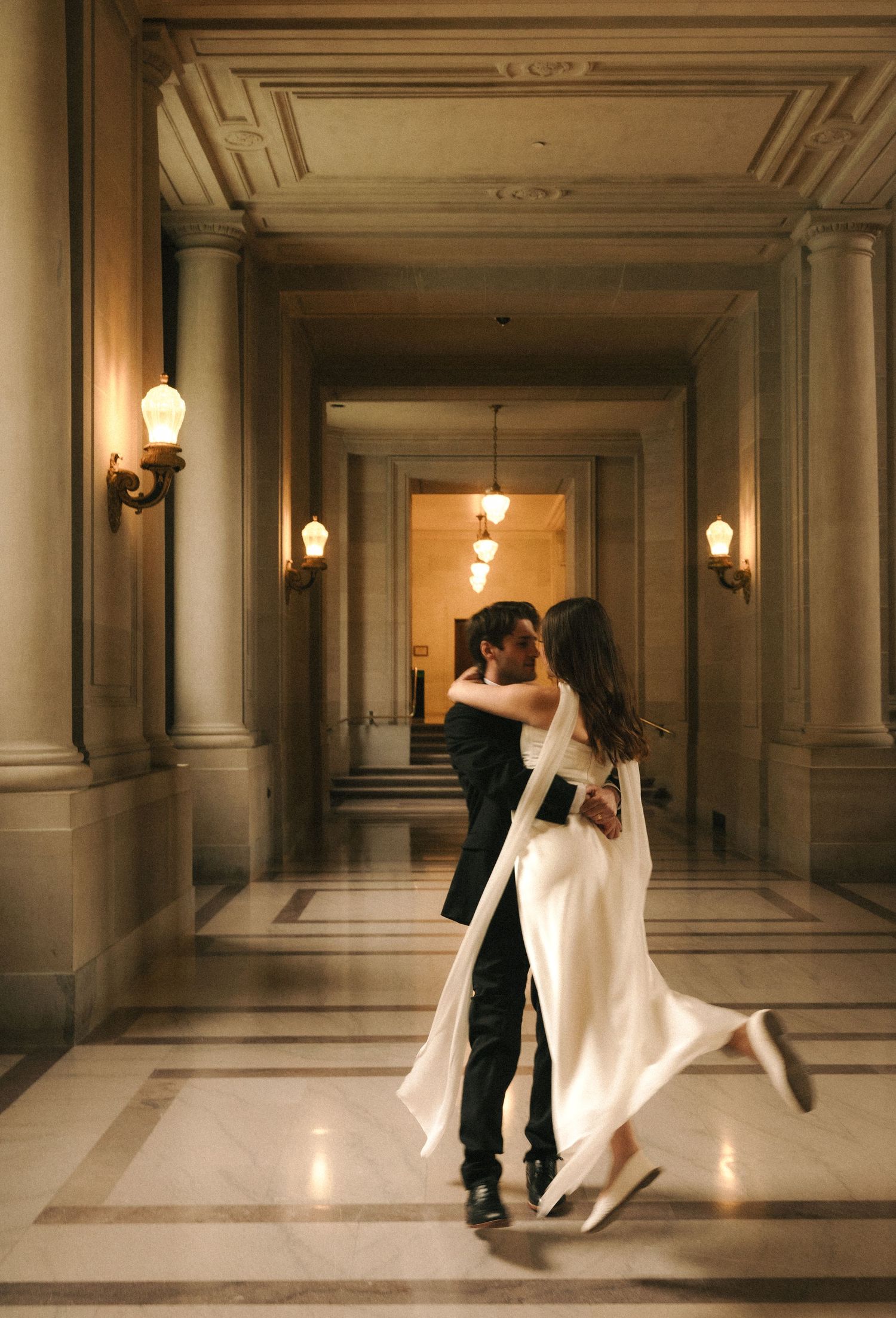 A couple shares a romantic kiss in an elegant marble hallway, the bride in a flowing white gown lifting her foot gracefully.