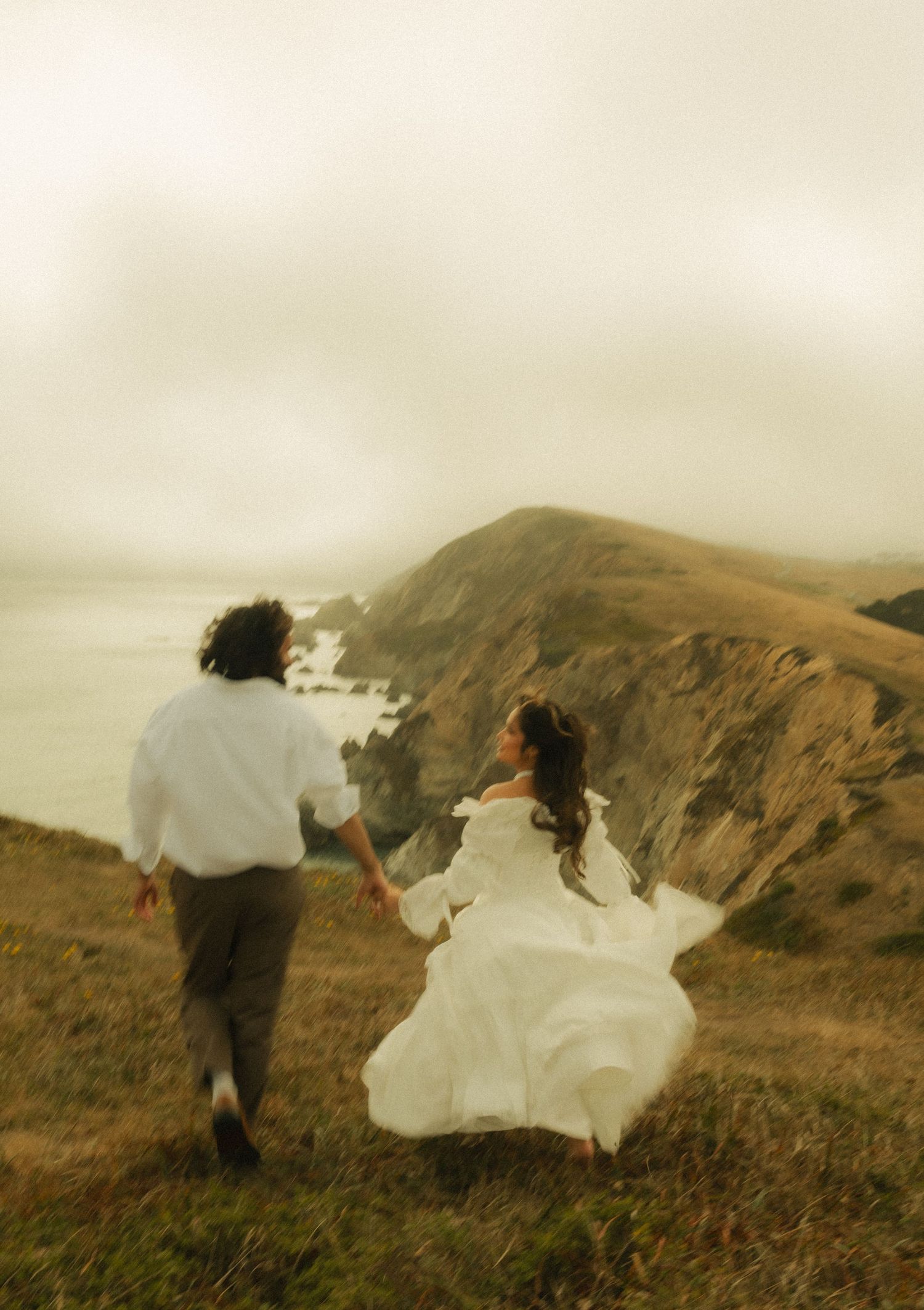 Bride and groom holding hands running on coastal cliffs in wedding attire, misty ocean backdrop.
