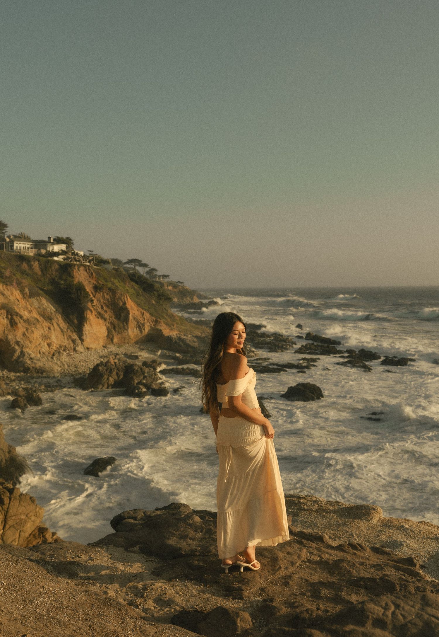 A woman in a white off-shoulder maxi dress stands on rocky coastal cliffs at golden hour with ocean waves behind her.