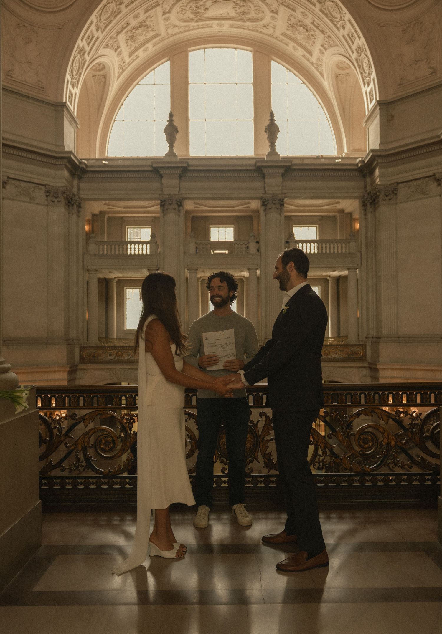 Couple exchanges vows at San Francisco City Hall with officiant, elegant Beaux-Arts architecture and grand arched window backdrop.