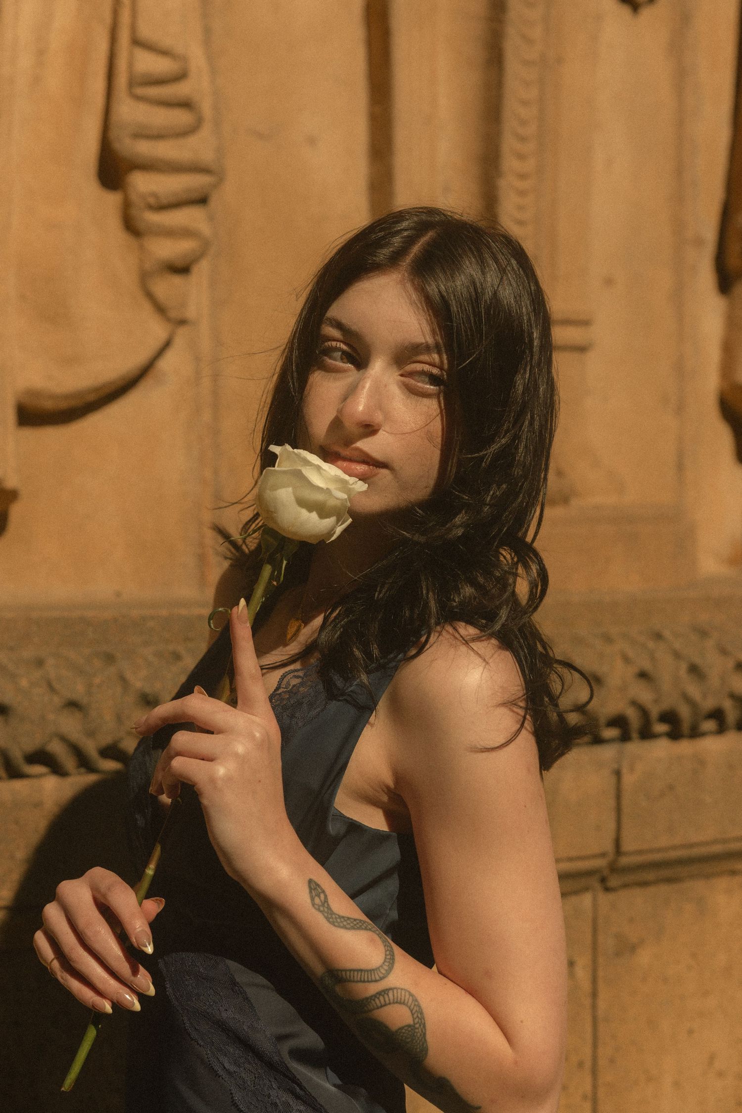 Young woman with dark hair holds a white rose, wearing a blue dress with a snake tattoo on her hand.
