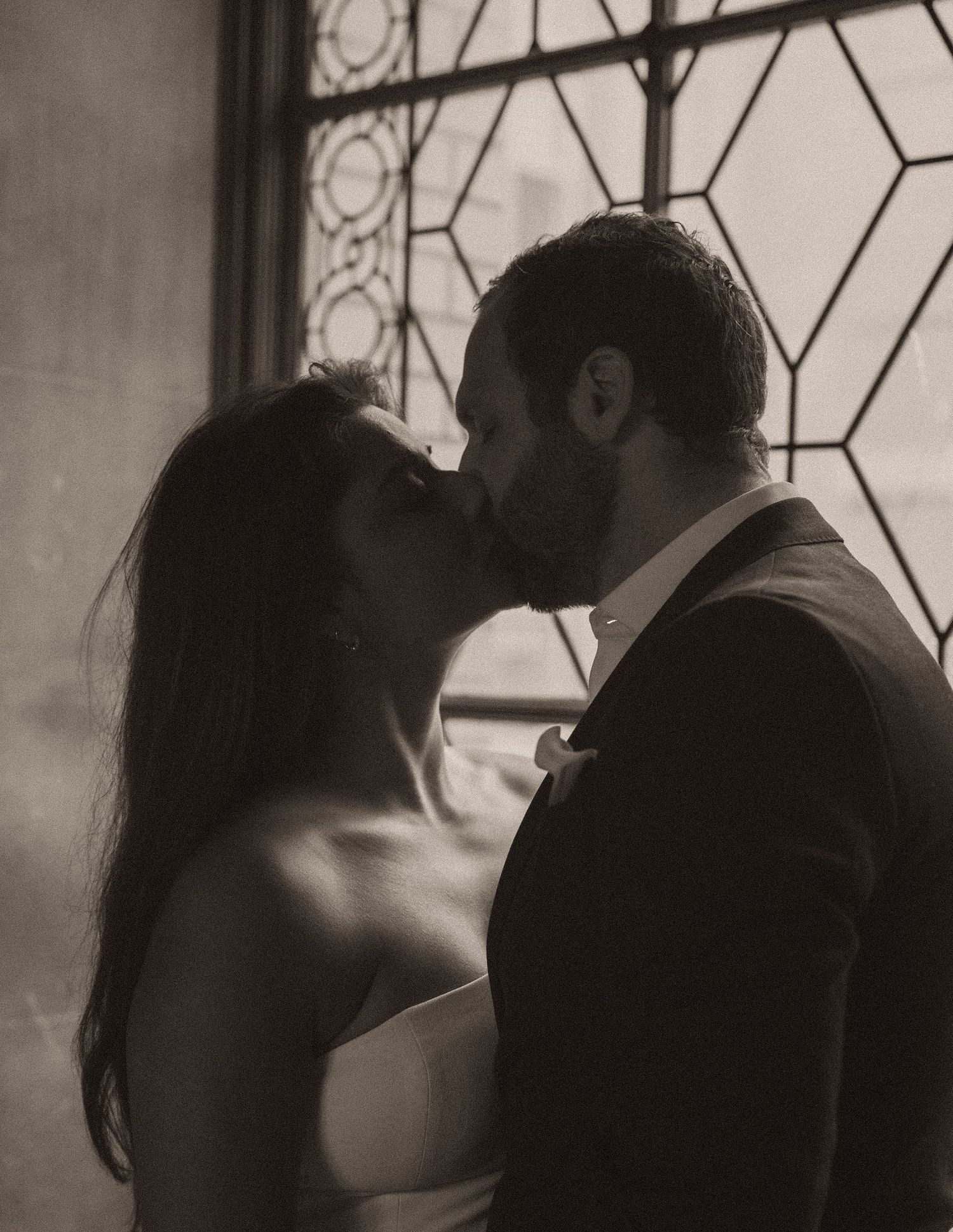 Couple sharing a romantic kiss in sepia tone, framed by a geometric stained glass window at their wedding.