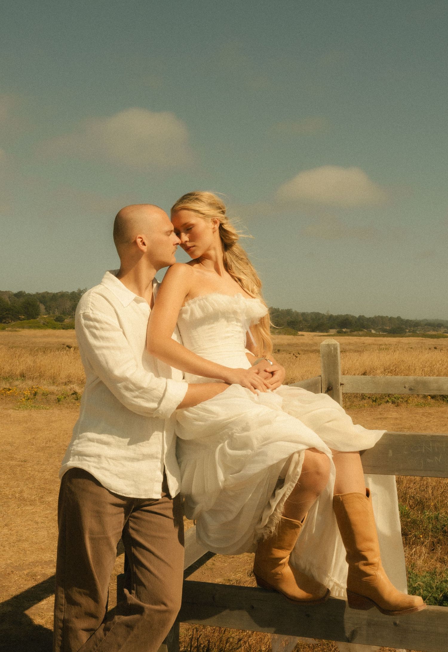 Romantic couple in wedding attire sitting on a fence in a golden field, bride in white dress and boots.