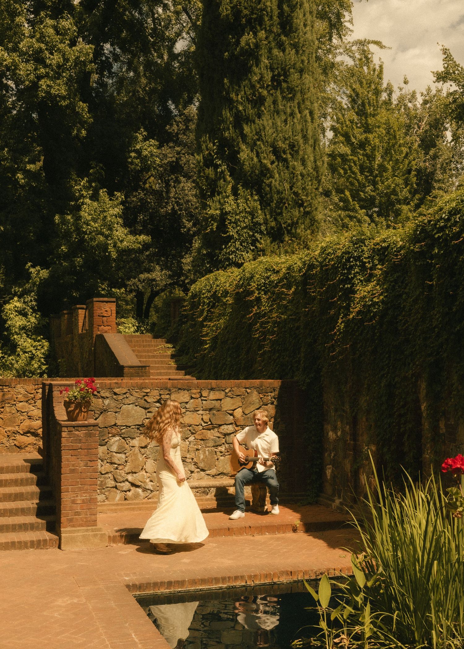 Man proposing to woman in elegant garden with stone walls, lush greenery, and reflecting pool on sunny day.