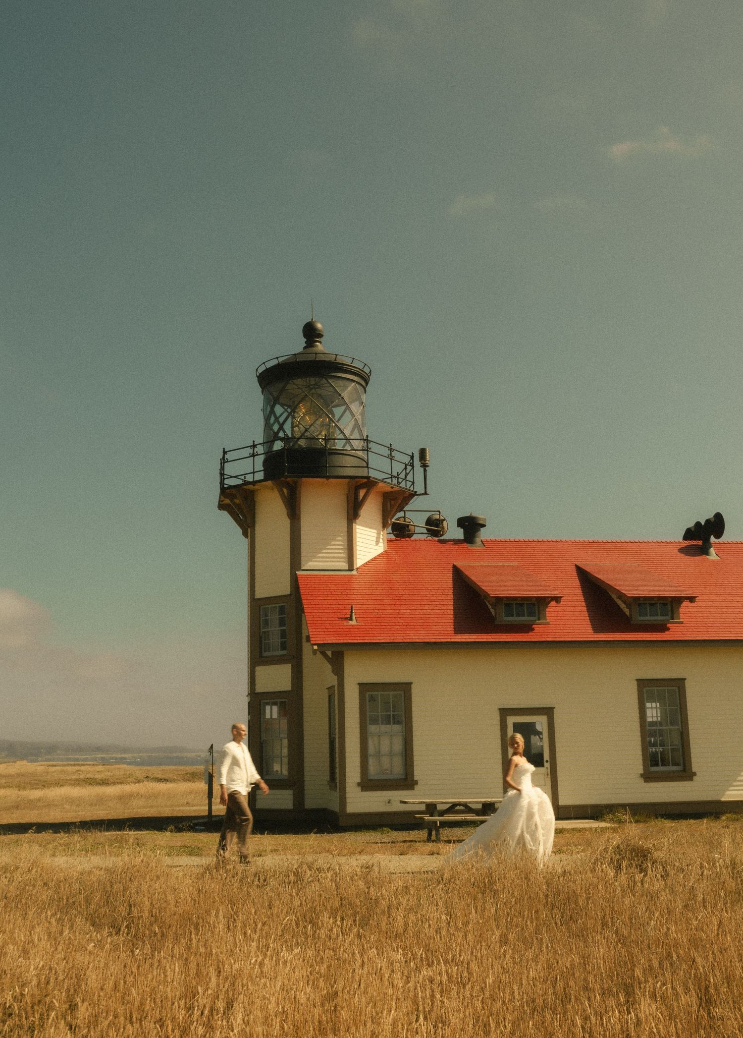 Bride and groom walk near a historic lighthouse with a red roof surrounded by golden fields under a blue sky.