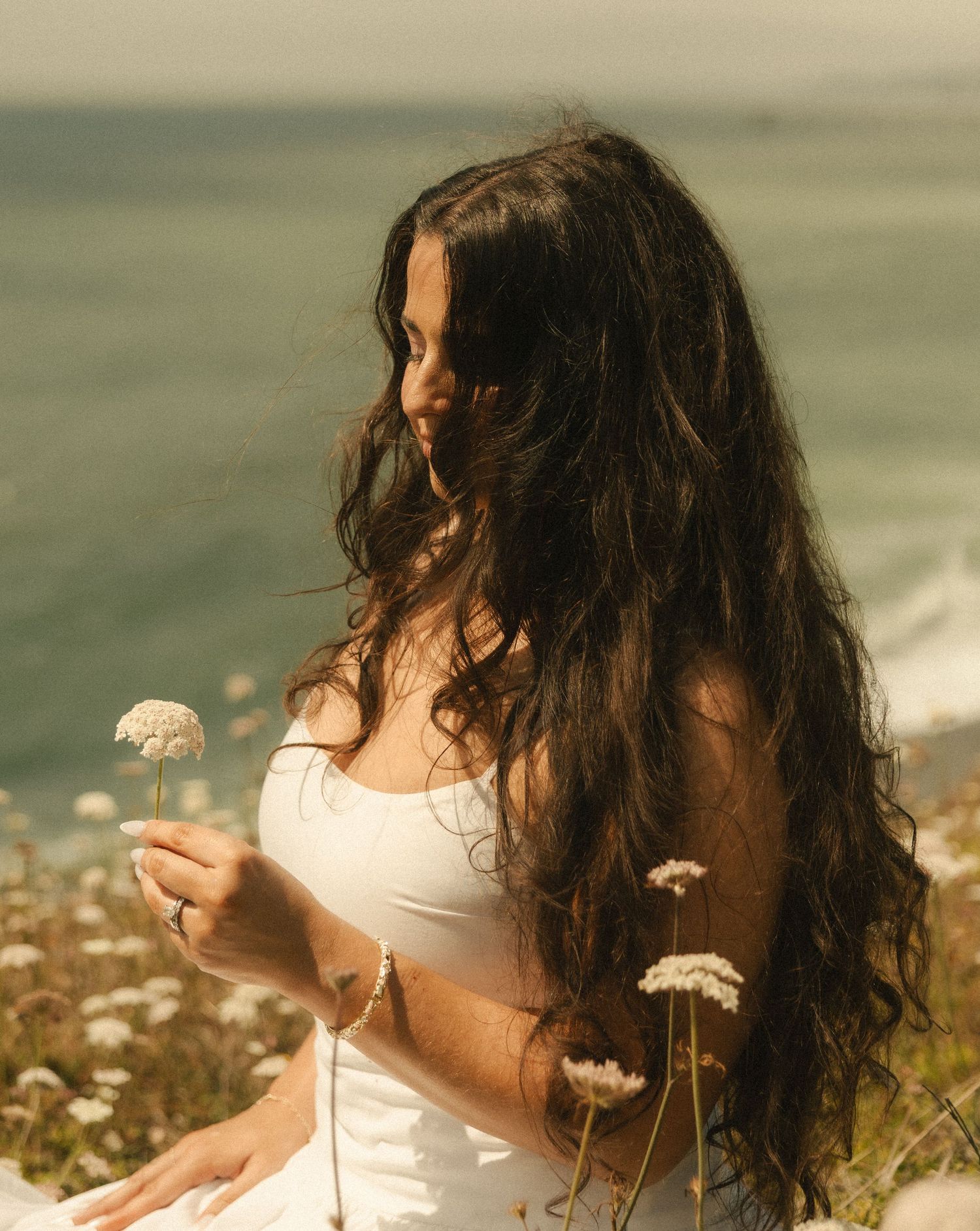 Woman with long dark wavy hair in white dress holding wildflowers by the ocean on a sunny day.