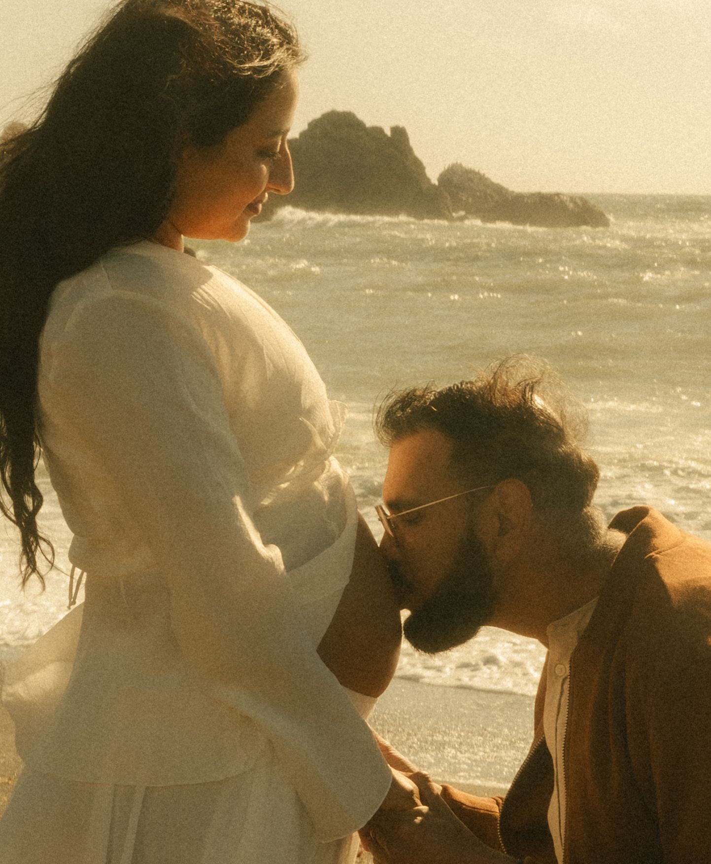 A romantic couple shares a tender moment on a beach, with the man kneeling to kiss the woman's hand.