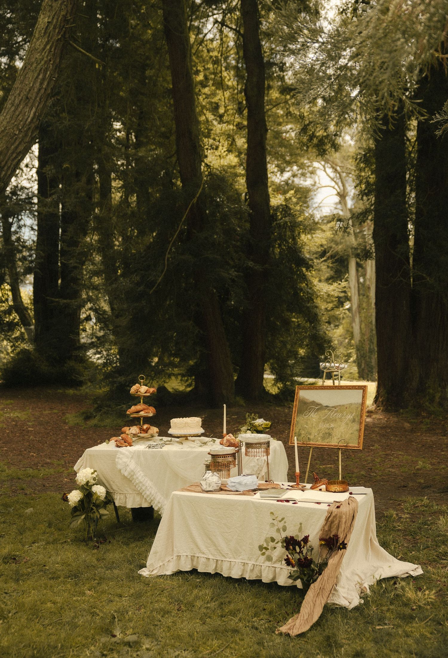 Elegant outdoor dessert table with white linen, floral arrangements, and a gold-framed painting set in a lush forest.