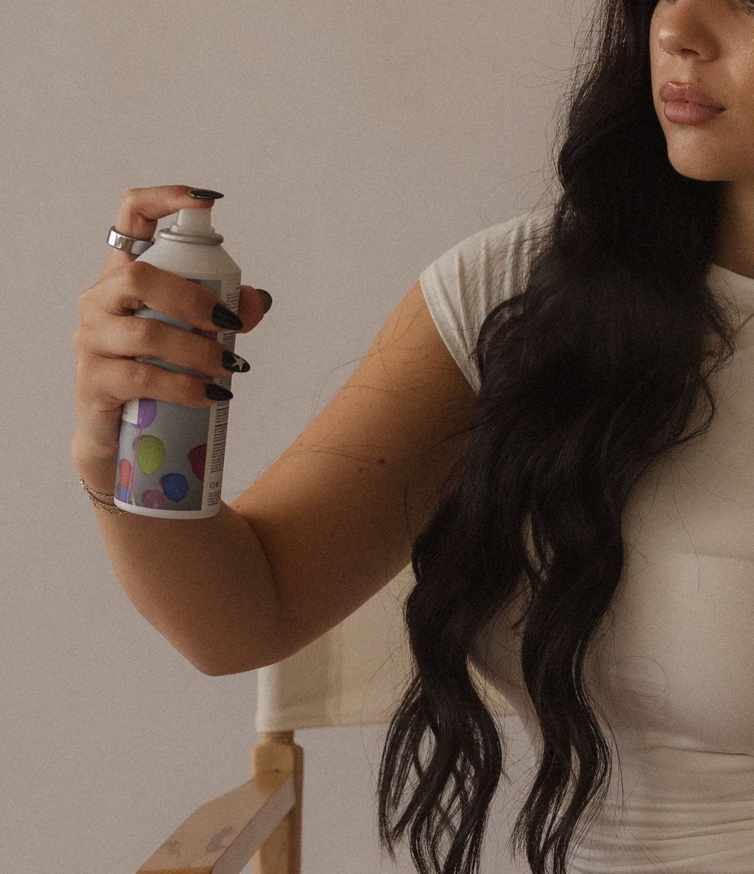 Woman with long dark wavy hair holding a colorful spray can hair product while wearing a white t-shirt.